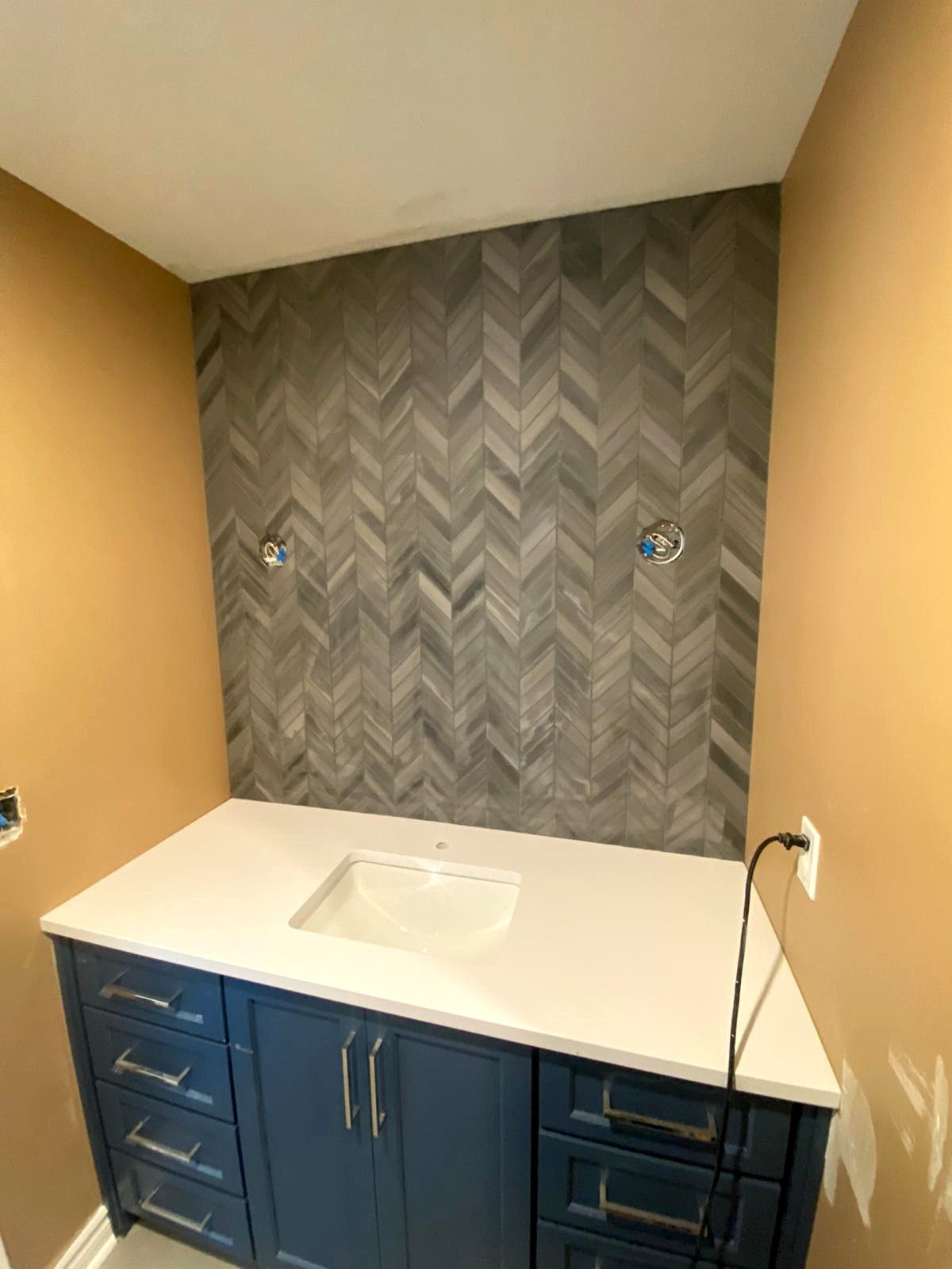 Bathroom vanity with blue cabinets, white countertop, and a space for a sink, against a wall with patterned tile, unfinished with electrical outlets and plumbing fixtures.