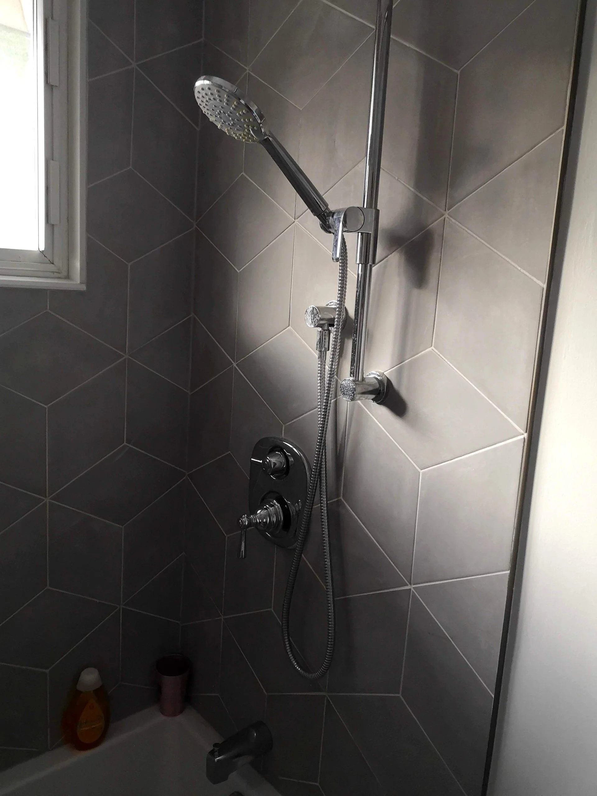 Shower with gray hexagonal tiles, a handheld showerhead, control knobs, a small window, and toiletries on the ledge.