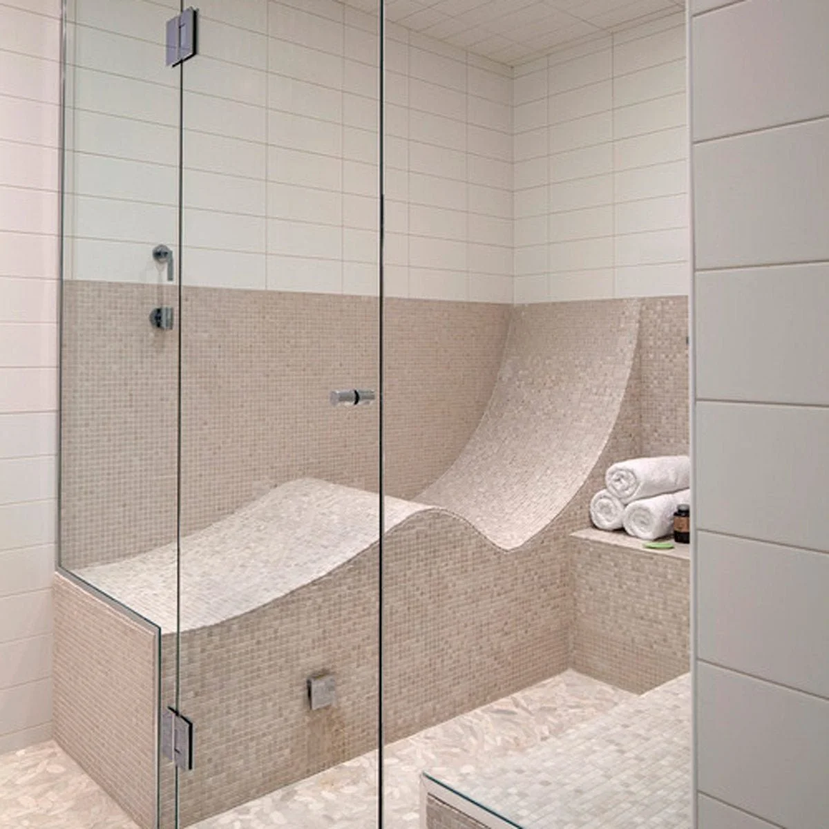 A modern shower area with a glass enclosure and beige mosaic tile bench featuring a wave-shaped surface for seating or bathing.