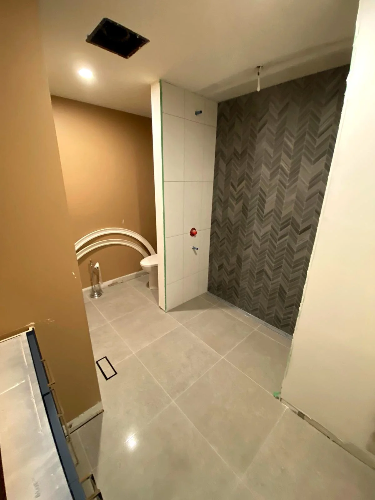 Undecorated bathroom with large beige tile flooring, a tan wall, a white toilet, partially tiled wall with white and gray patterned tiles, ceiling with cutout for lighting, and construction materials present.