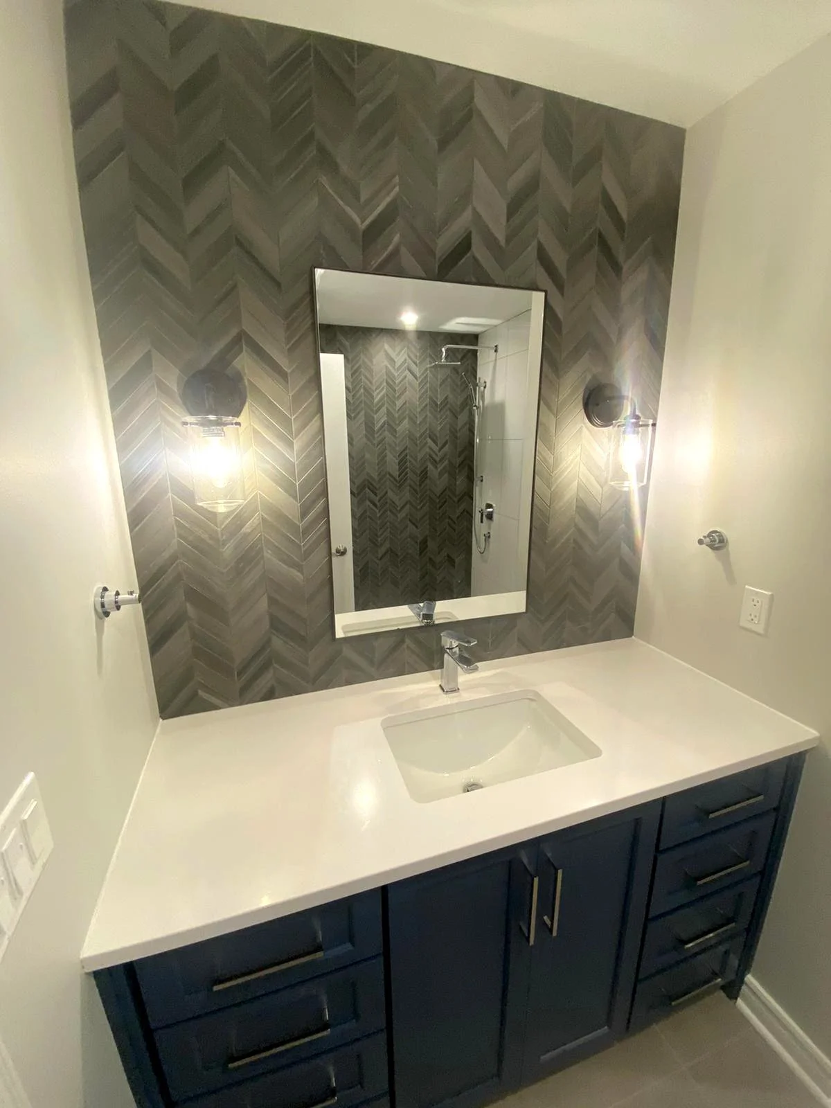 Bathroom vanity with a white countertop, a built-in sink, dark blue cabinets, a large mirror, wall-mounted light fixtures, and a patterned accent wall, with visible shower area in reflection.