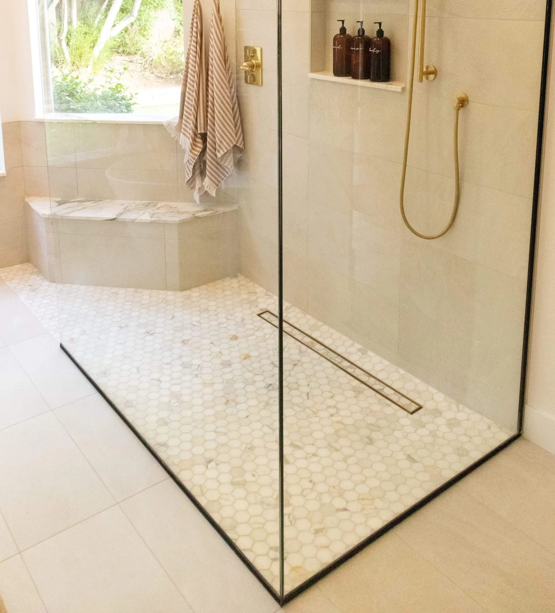 Modern walk-in shower with glass enclosure, beige hexagonal floor tiles, a wall-mounted shelf with three soap dispensers, a handheld showerhead, and a built-in bench near a window with striped towels hanging.