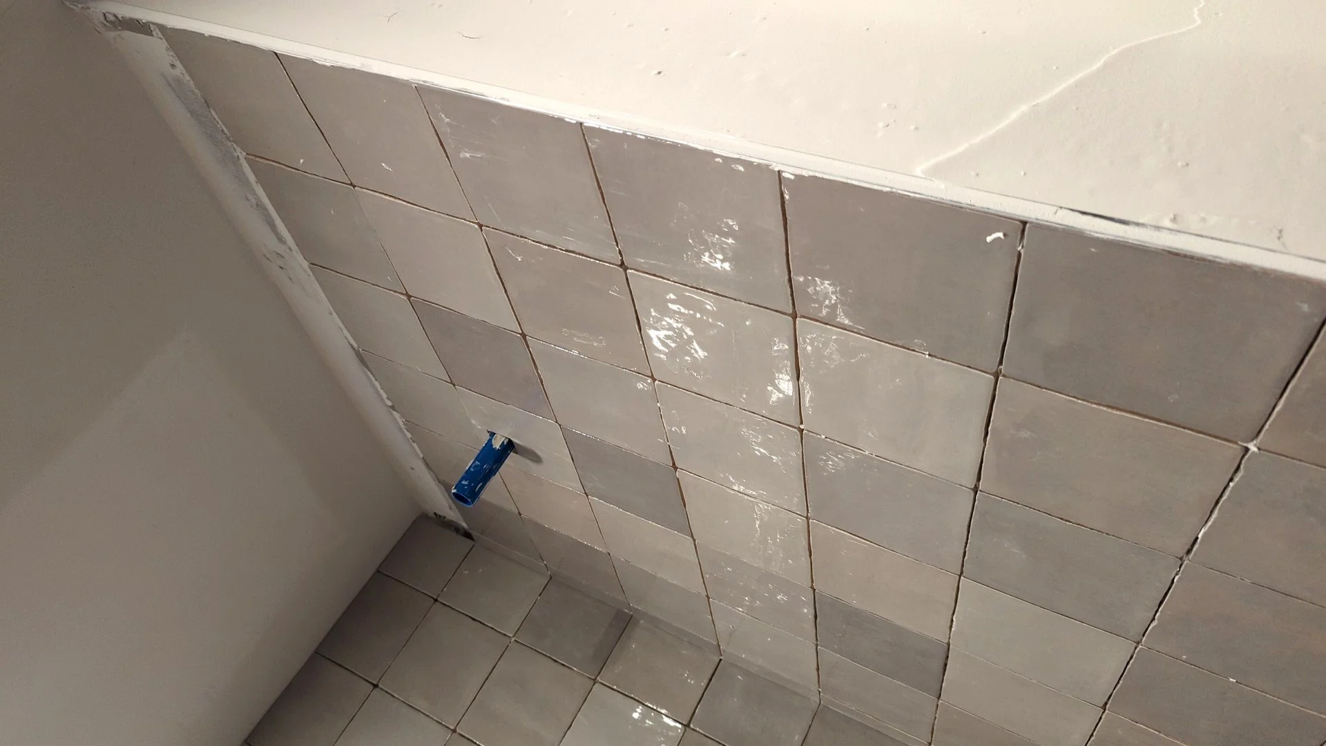 Close-up of a tiled wall and floor in a bathroom, showing a partially installed toilet tank with a blue flush pipe.