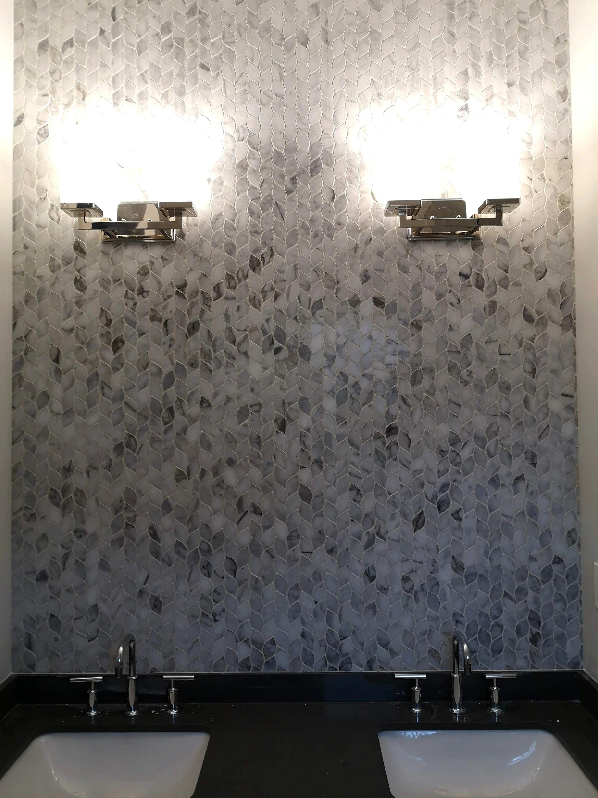 Bathroom with a textured grey tile wall, two wall-mounted lights, and two sinks with black countertops.