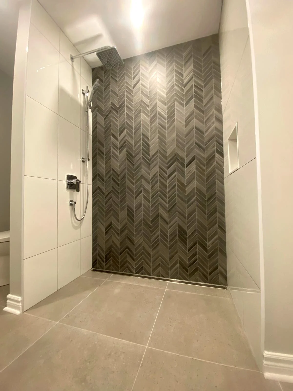 Modern shower with a square rainfall showerhead and a hand-held shower, tiled with chevron pattern dark gray tiles inside the shower area, and large beige tiles outside.