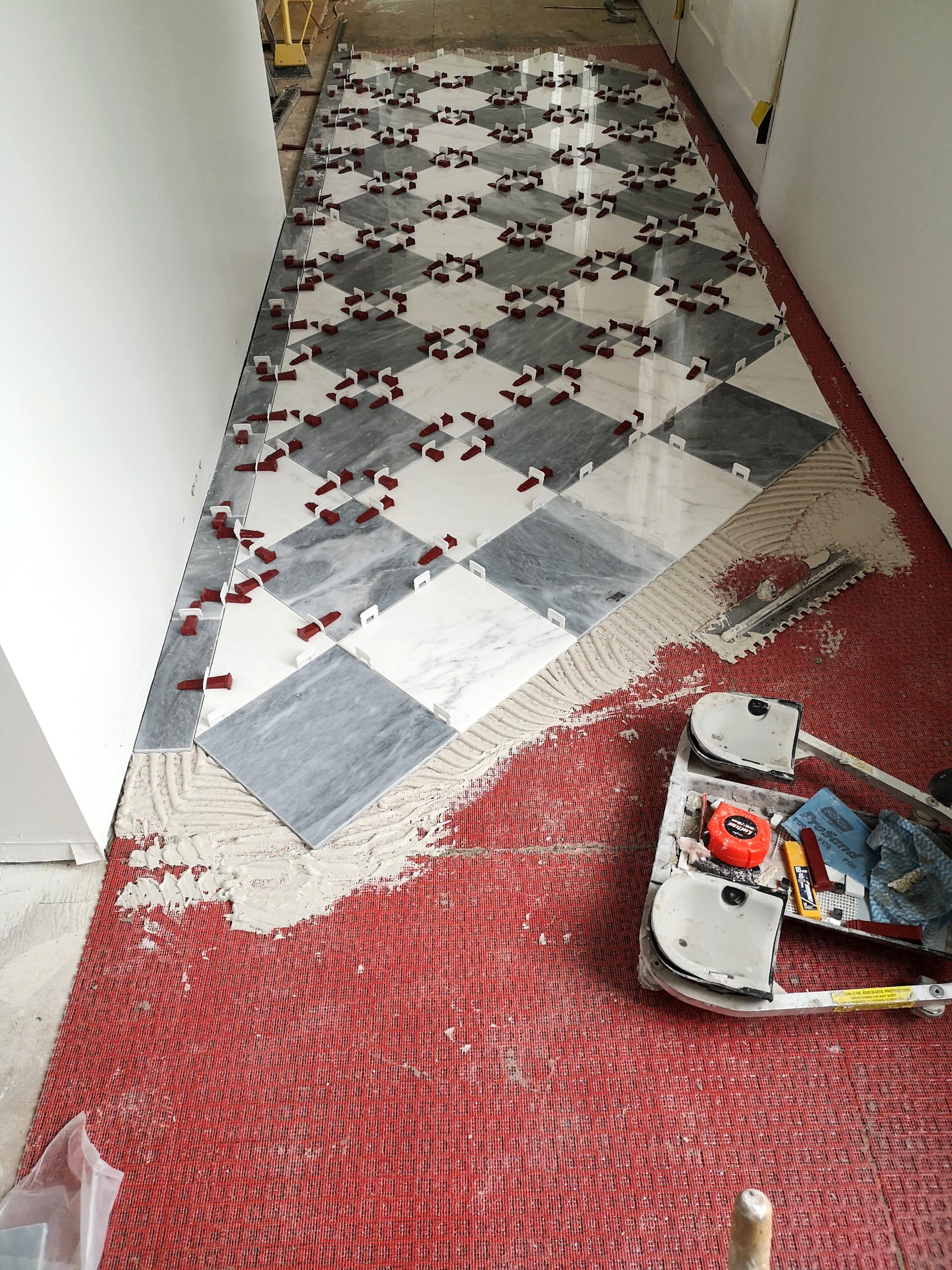 Inside a building under renovation, new marble tile flooring with a black and white pattern is being installed. The tiles are partially laid, and tile spacers are visible between them. Red carpet protection is laid out on the floor, with some areas r