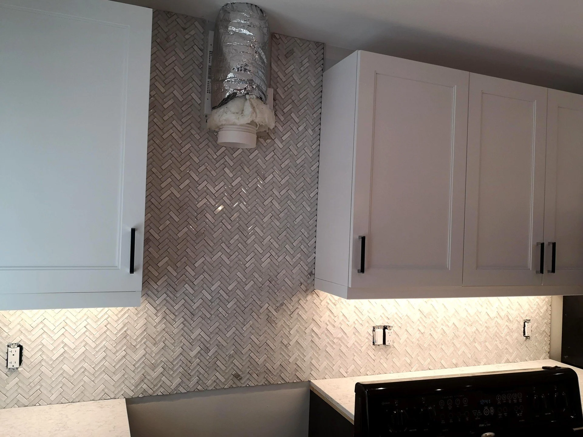 Kitchen backsplash with herringbone tile pattern, white upper cabinets with black handles, electrical outlets, and a vent with insulation, under-construction or renovation.