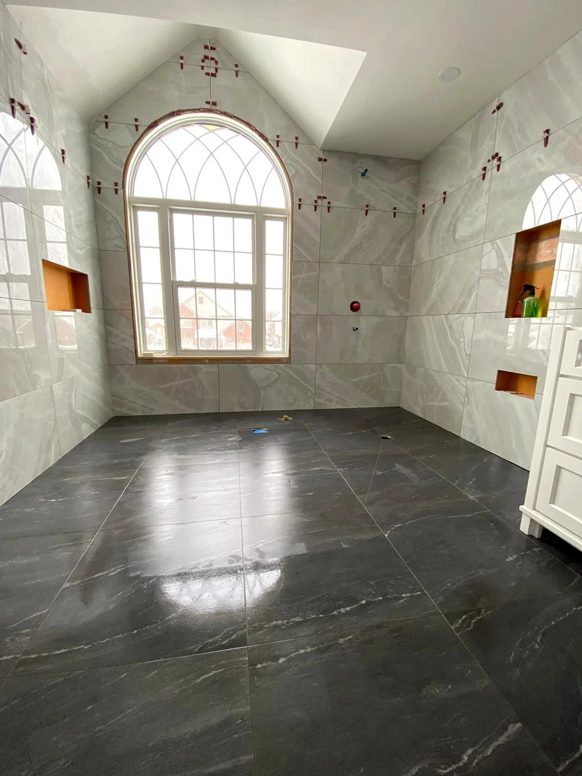 An unfinished bathroom with large black floor tiles, marble wall tiles, and a large arched window with a grid pattern, letting in natural light.