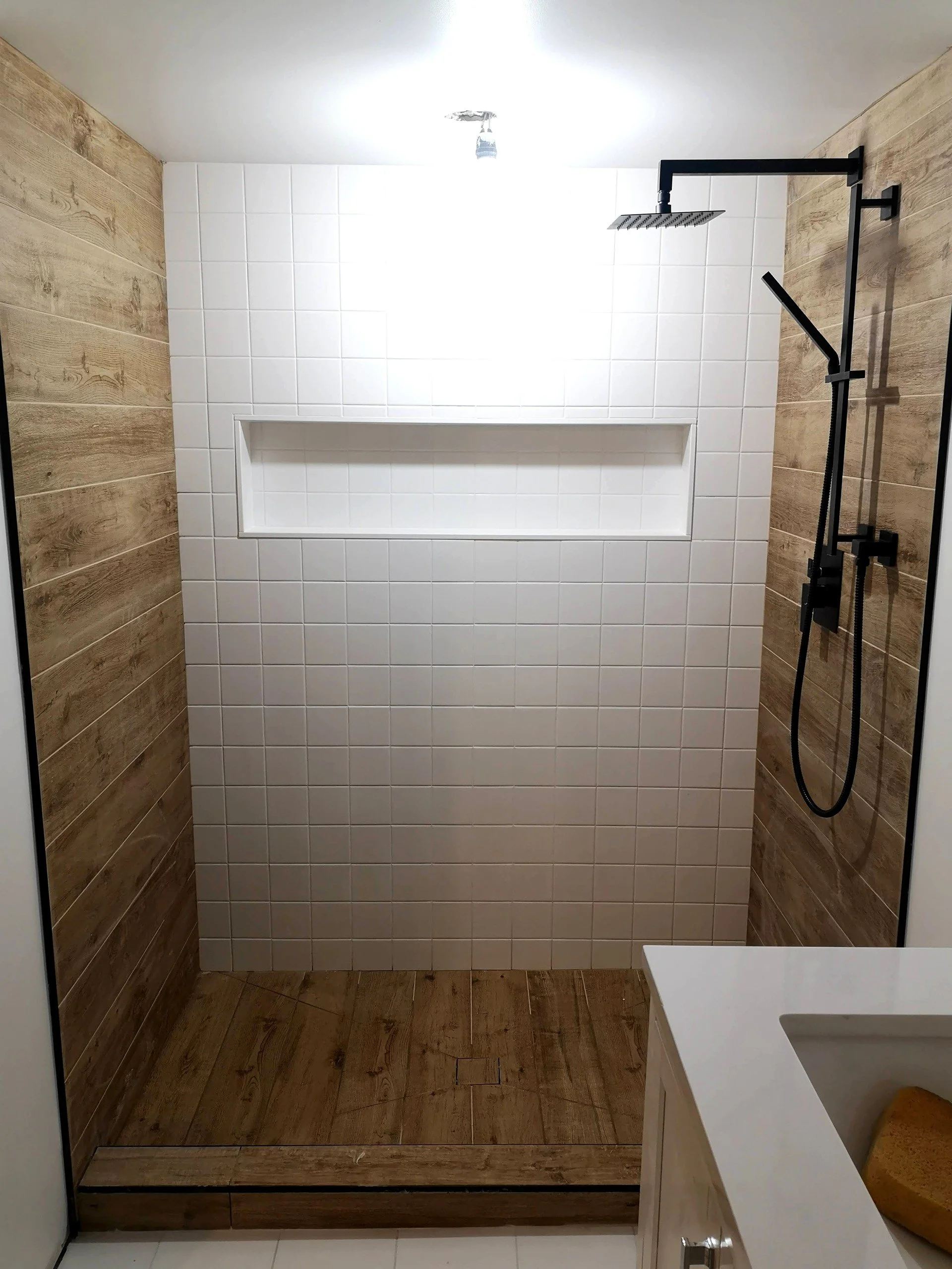 Empty modern shower with white tiled wall, wooden wall panels, black rain showerhead and handheld shower, and a small built-in shelf in the tiled wall.