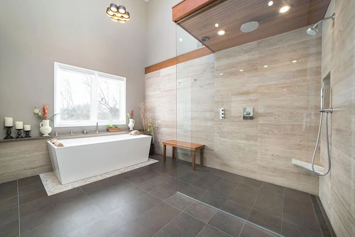 Modern bathroom with a large walk-in shower on the right, featuring beige tiled walls and silver fixtures. On the left, there's a bathtub beneath a window, decorated with candles and vases with flowers. The bathroom has a high ceiling with a contemporary light fixture and natural light coming in through the window.