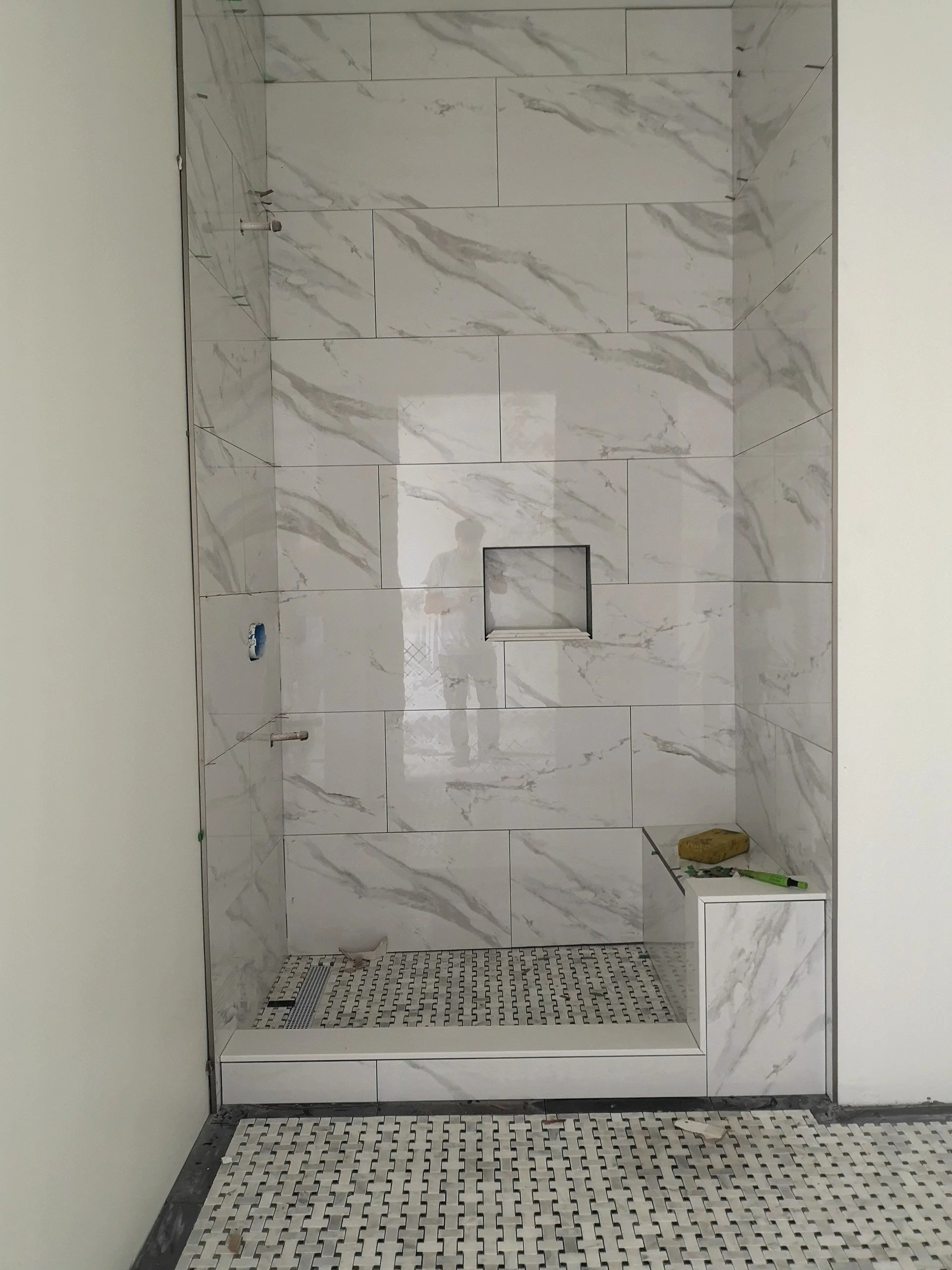 Newly tiled walk-in shower with marble-like wall tiles, a niche in the wall for toiletries, and a small ledge with construction tools. The shower floor has small patterned tiles, and some tools and construction debris are visible.