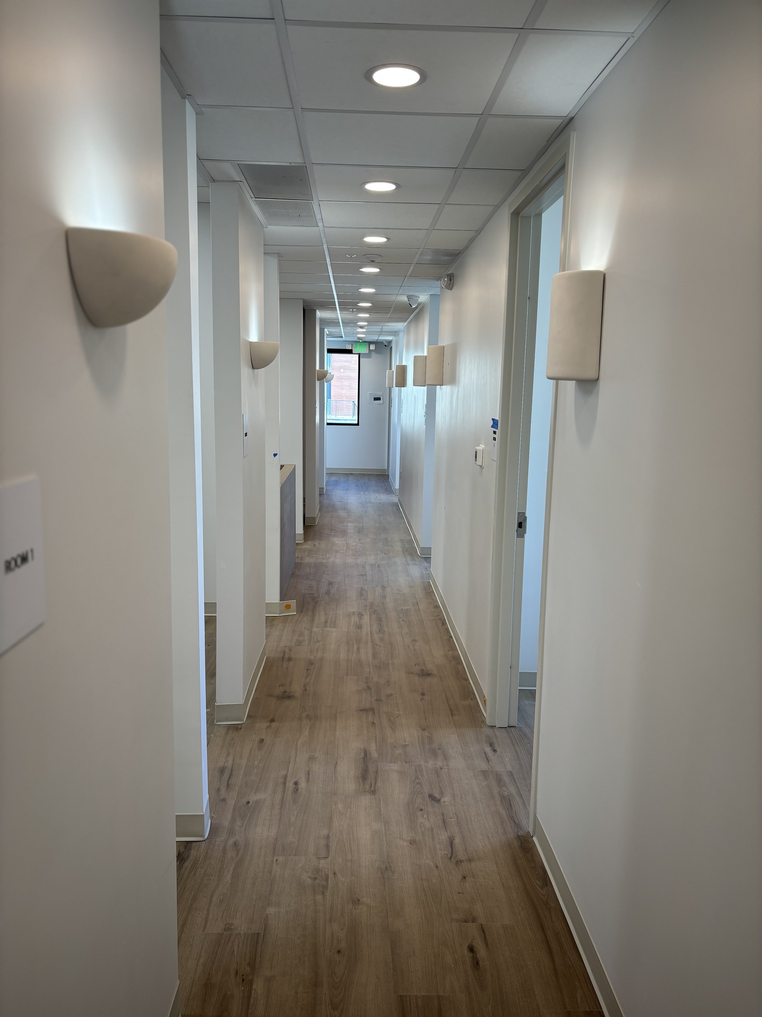 Hallway with wood looking LVP/luxury vinyl plank flooring and white painted walls, featuring wall-mounted electrical lights and a window at the end. Your go to for 3rd party construction takeoffs