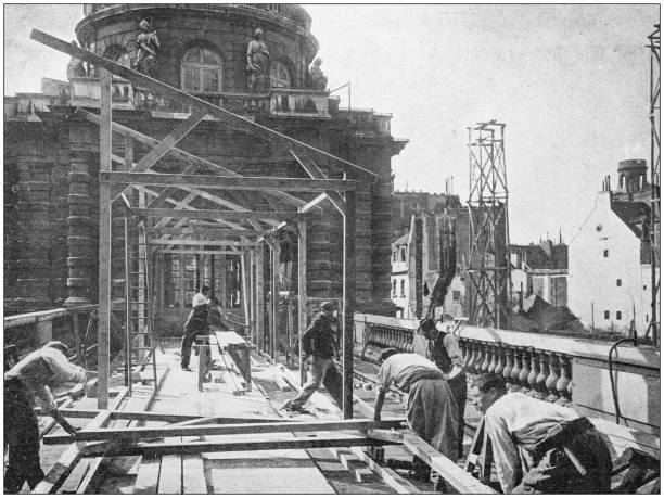 Black and white photo of commercial construction workers building a structure on a city street. The specialty contractors or subcontractors are seen working with wood, and a large ornate building with statues and windows is visible in the background.
