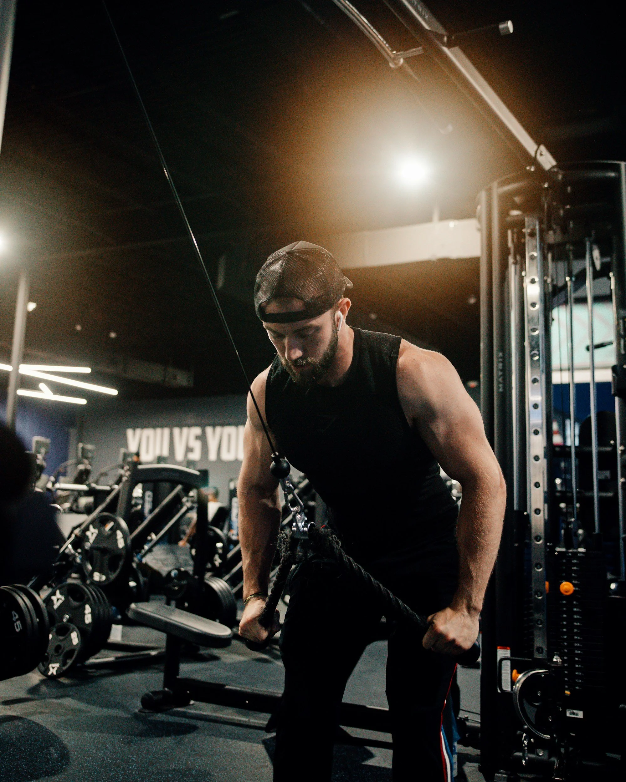A man in a black sleeveless shirt and cap working out with a cable machine at a gym. The gym has various exercise equipment and a motivational quote on the wall that says 'YOU VS YOU'.