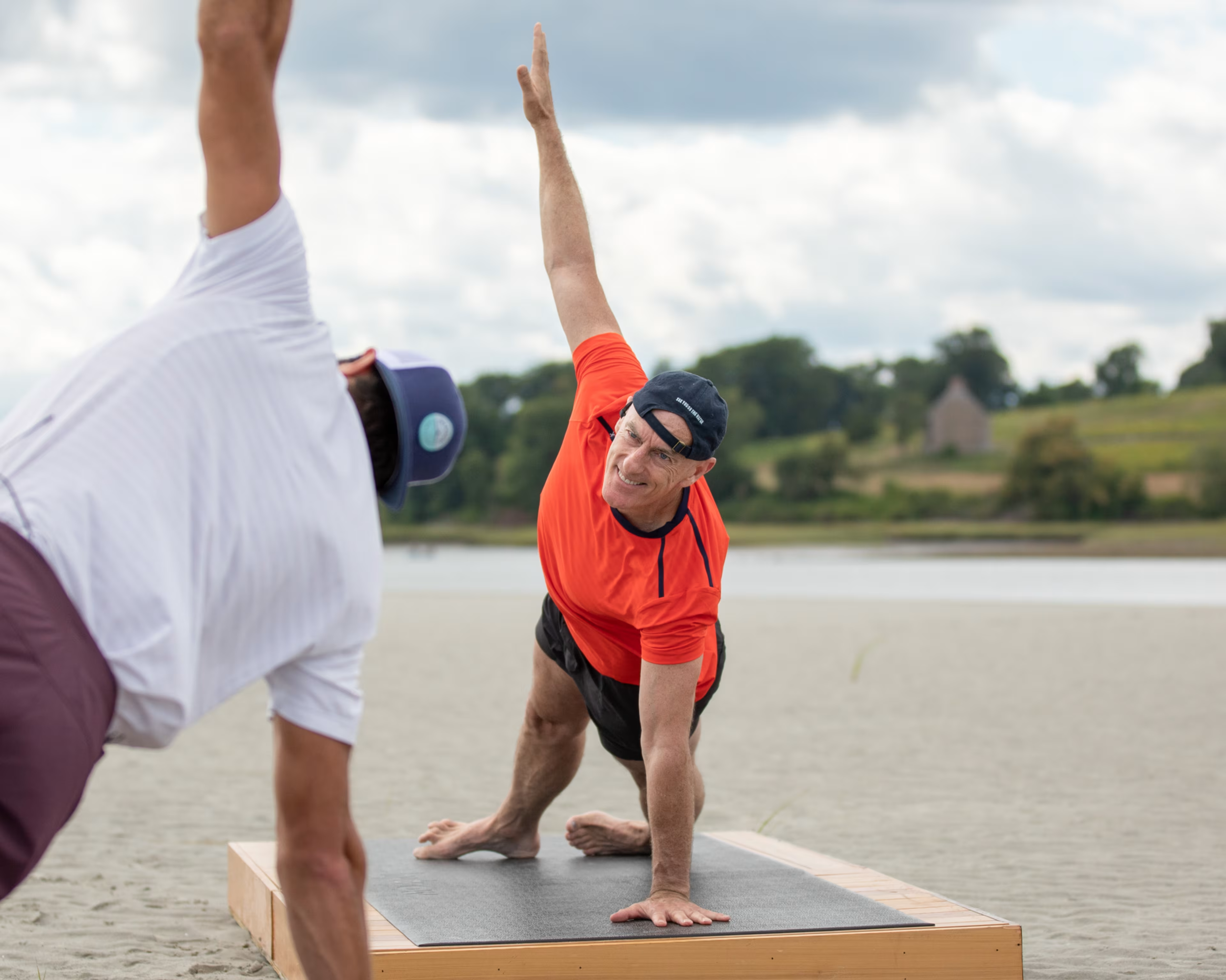 Two men practicing yoga outdoors on a beach, one in an orange shirt and black shorts, performing a side plank pose, while the other in a white shirt and maroon shorts observes.
