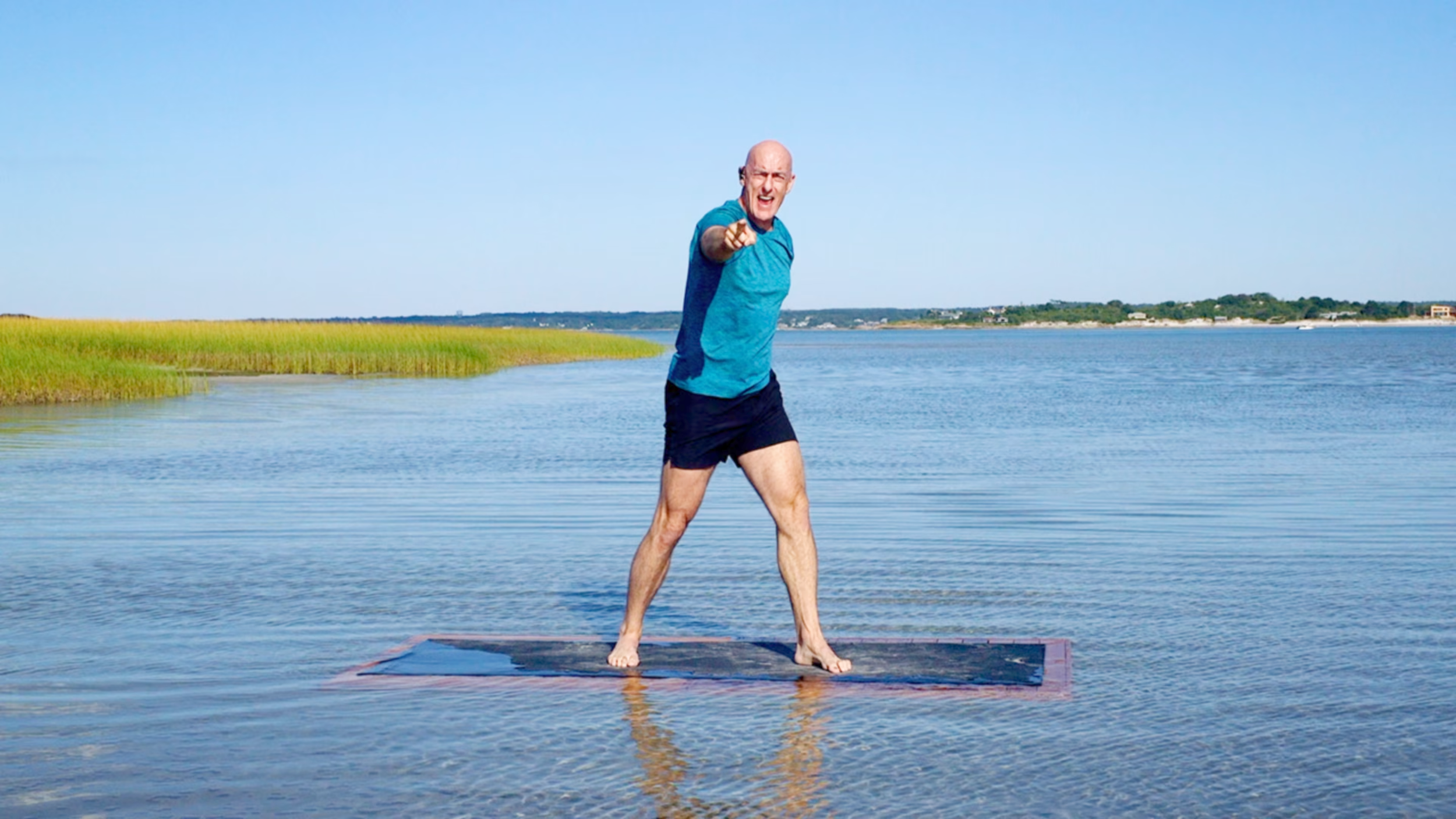 A man in a blue shirt and black shorts standing barefoot on a floating yoga mat near the water, pointing towards the camera with a smiling face on a sunny day.
