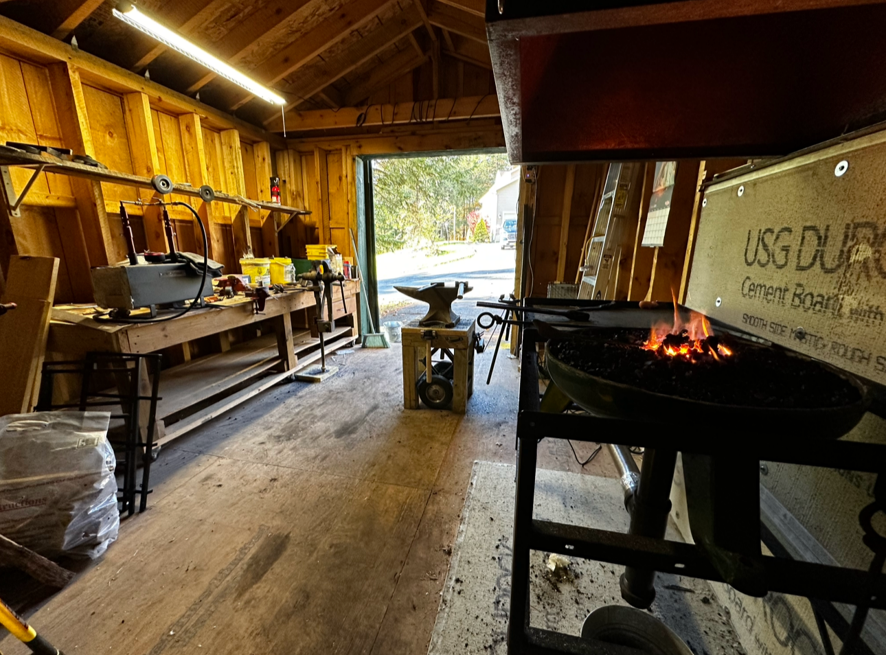 This is a woodworking workshop with tools and equipment, including a workbench, a vise, and a forge with a fire. The space has wooden walls and a large open door leading outside.