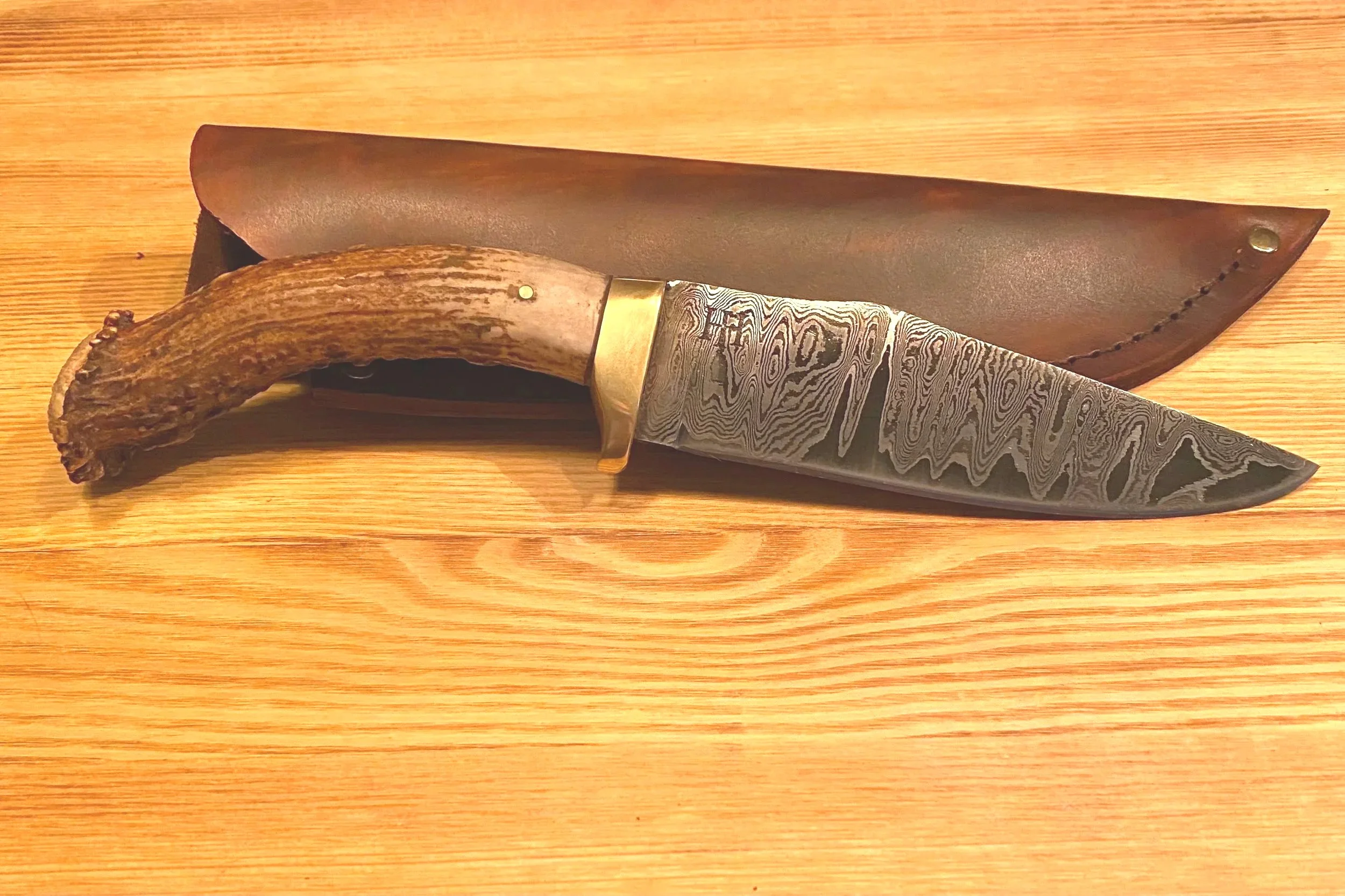 A folding knife with a patterned blade, a brass guard, and a handle made from a deer antler, resting on a wooden surface with a leather sheath behind it.