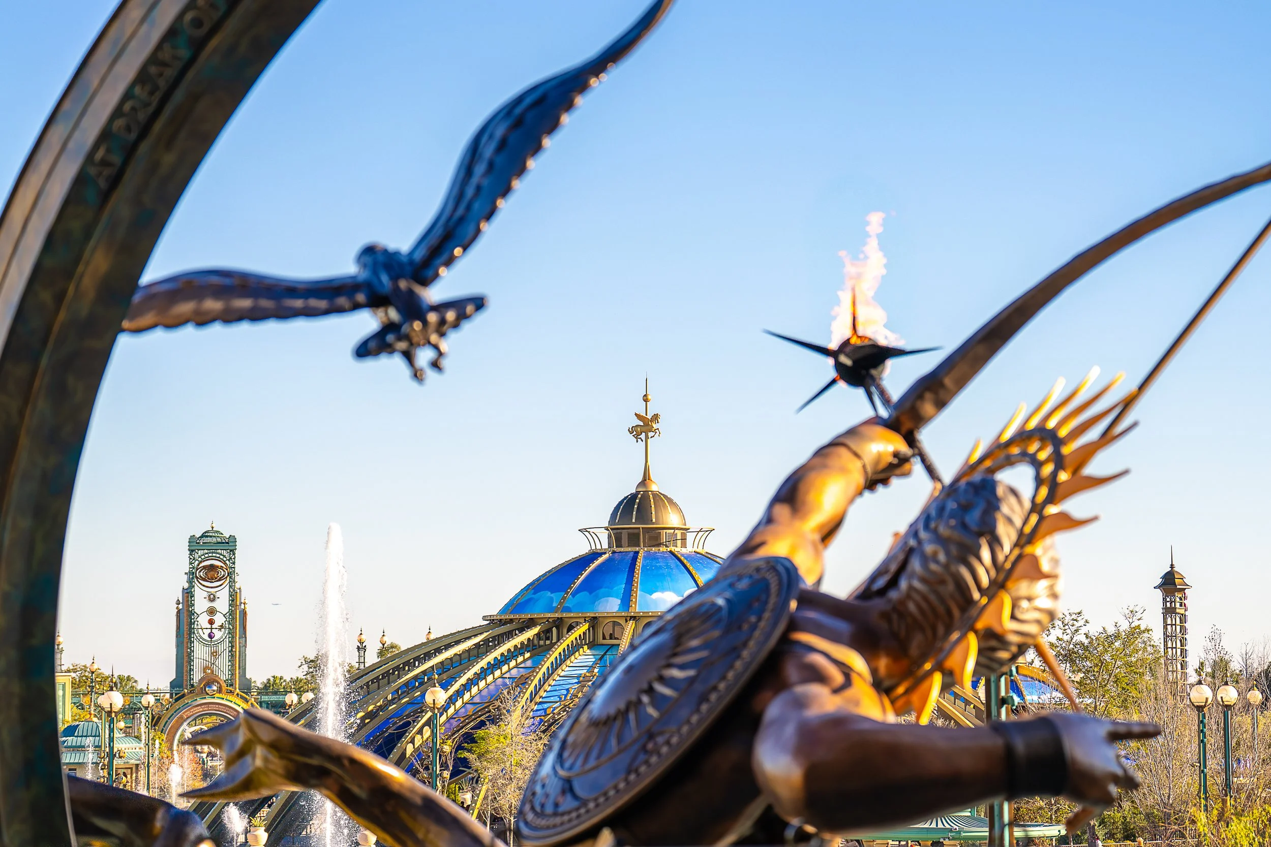 Epcot theme park with a sculpture of a person reaching, surrounded by dragonflies in the foreground, and the park’s pavilion with a blue domed roof and spire in the background.