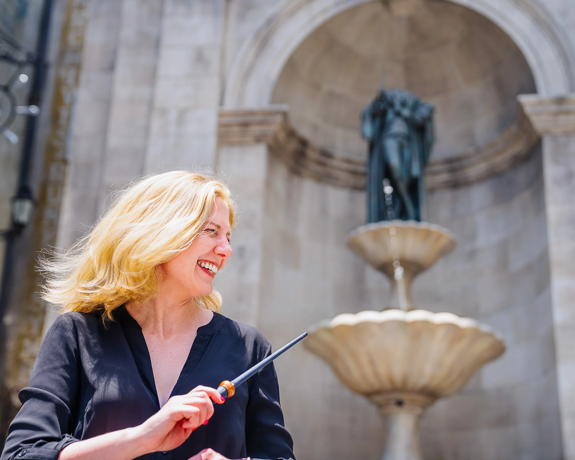 Smiling woman with blonde hair holding a wine opener in front of a stone fountain and statue.