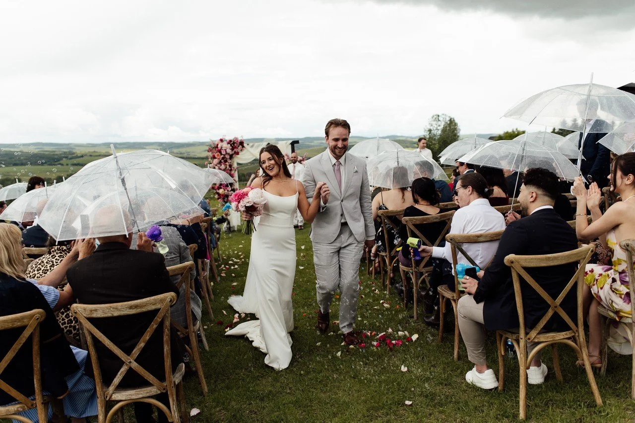 A bride and groom walking down an outdoor wedding aisle after their ceremony, holding hands and smiling. The bride is holding a bouquet of pink and white flowers, and guests seated on either side are holding clear umbrellas, celebrating despite the rainy weather.