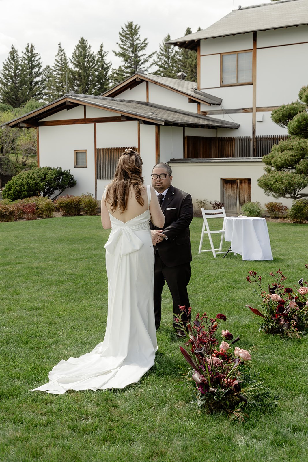 A bride and groom standing on a lawn with a house in the background during a wedding ceremony. The bride is wearing a white gown with a large bow at the back, and the groom is in a black suit with glasses. There are flowers arranged on the grass and a small table with chairs nearby.