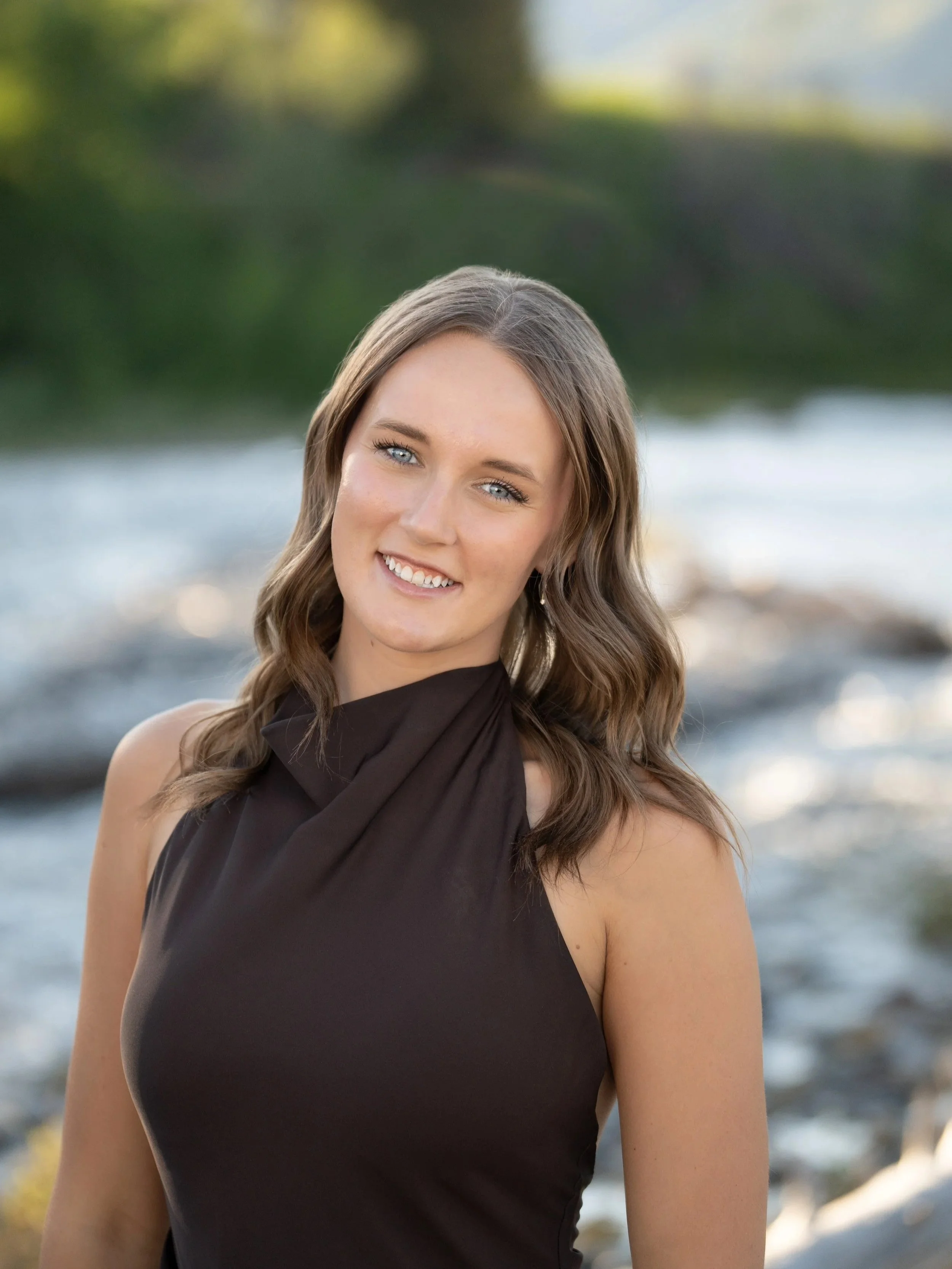 A woman with long brown hair and blue eyes smiling outdoors on a sunny day, wearing a sleeveless black top, with trees and water in the background.