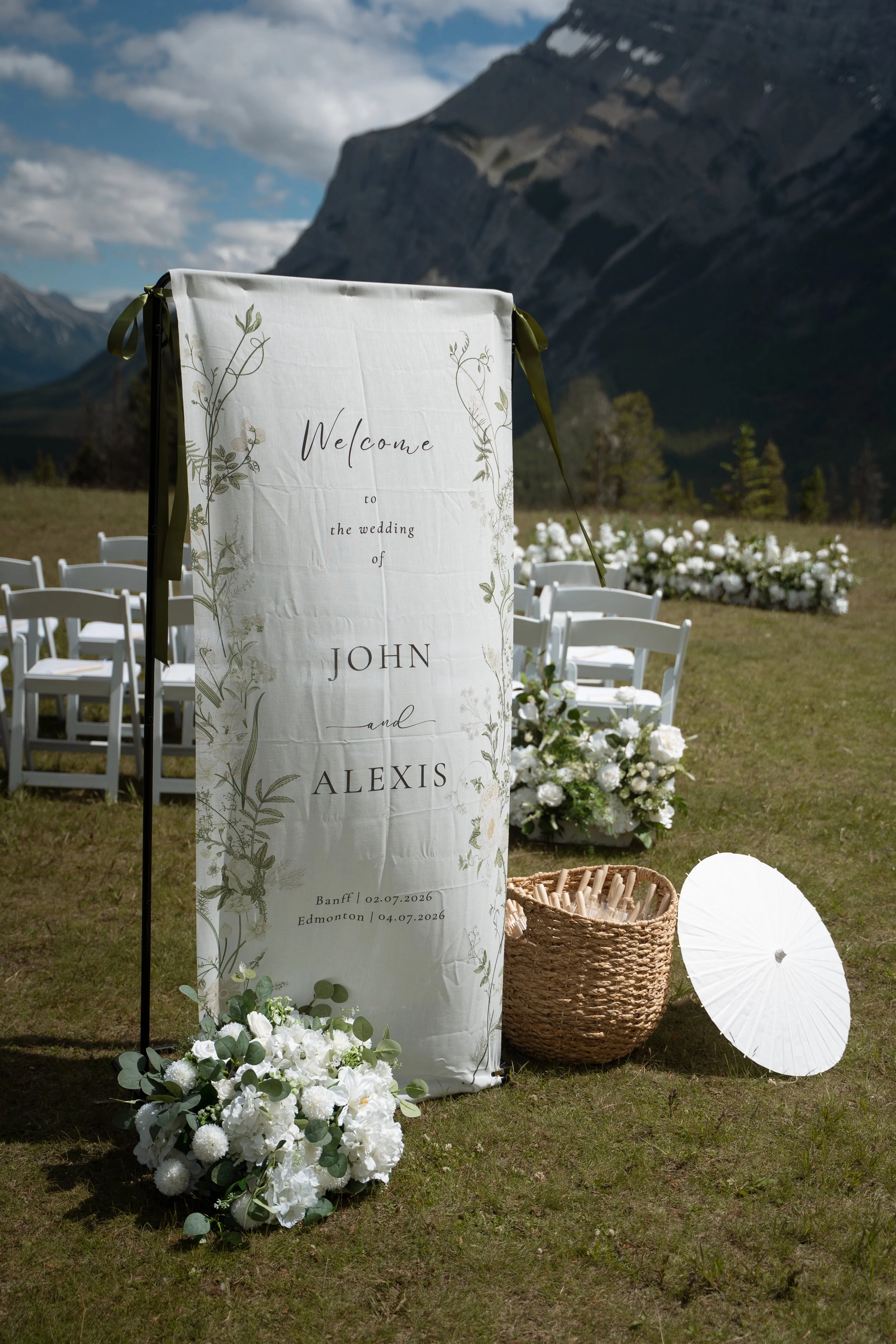 Outdoor wedding setup with a vertical welcome sign featuring floral designs and the names John and Alexis, set against mountains. Decor includes white flowers, a wicker basket, a white parasol, and white chairs.