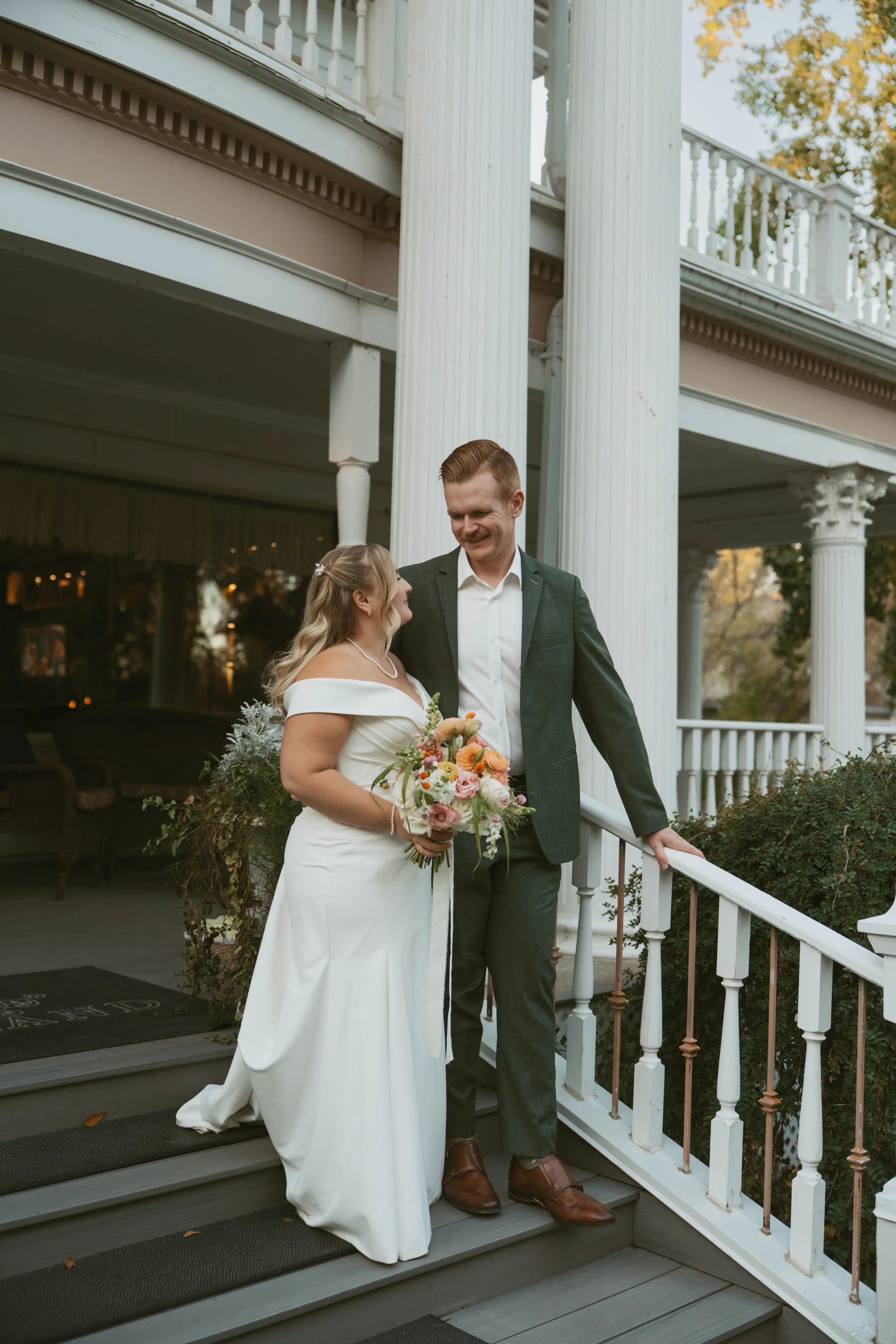 A bride and groom standing on a porch, smiling at each other. The bride is holding a bouquet of flowers and wearing a white wedding dress with off-the-shoulder sleeves. The groom is dressed in a dark suit with a white shirt. There are white columns and a railing on the porch, with trees in the background.