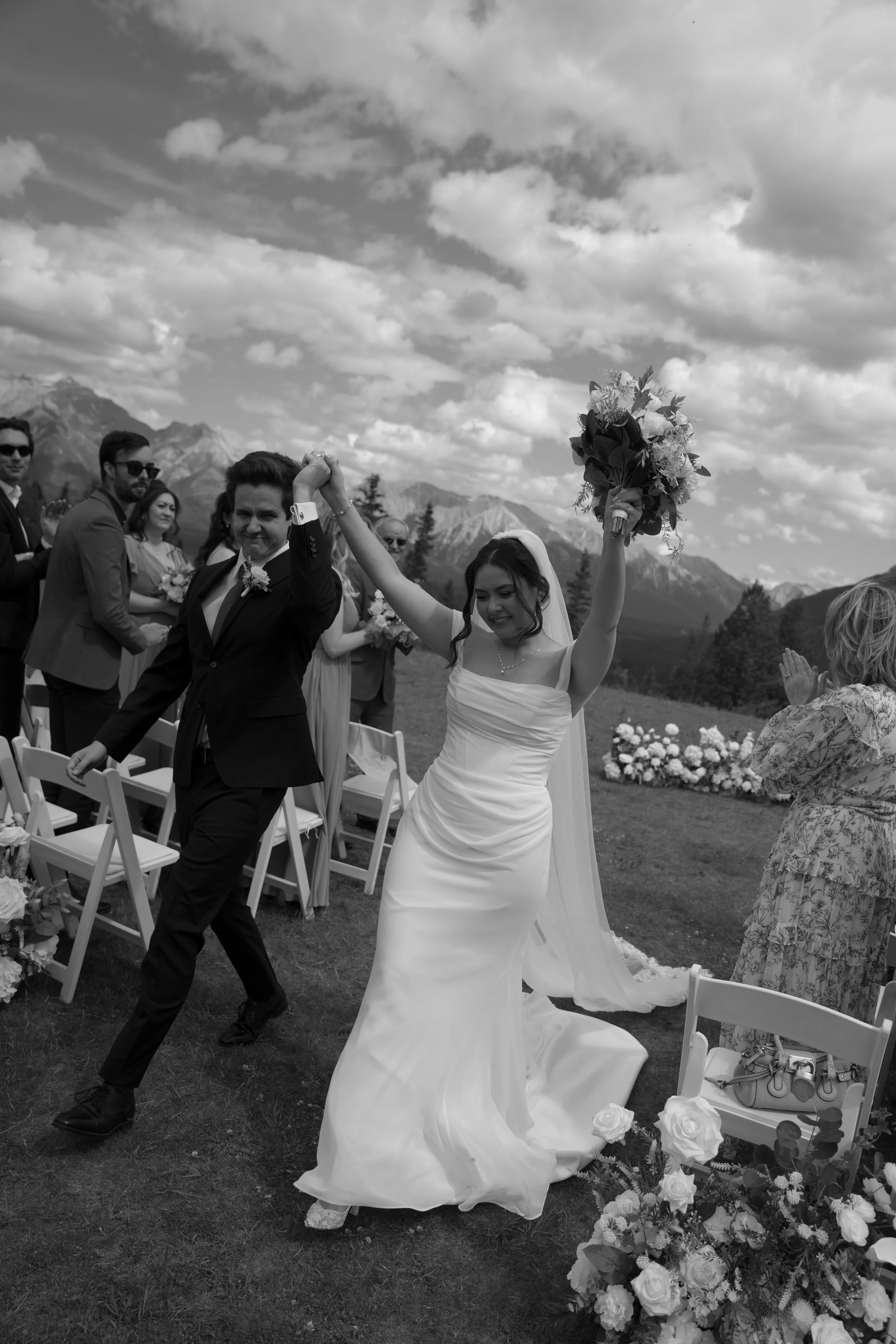 A black and white photo of a wedding ceremony outdoors. The bride, holding a bouquet, celebrates as she walks down the aisle with a groom, both smiling. Guests are seated on either side, with mountains and clouds in the background.