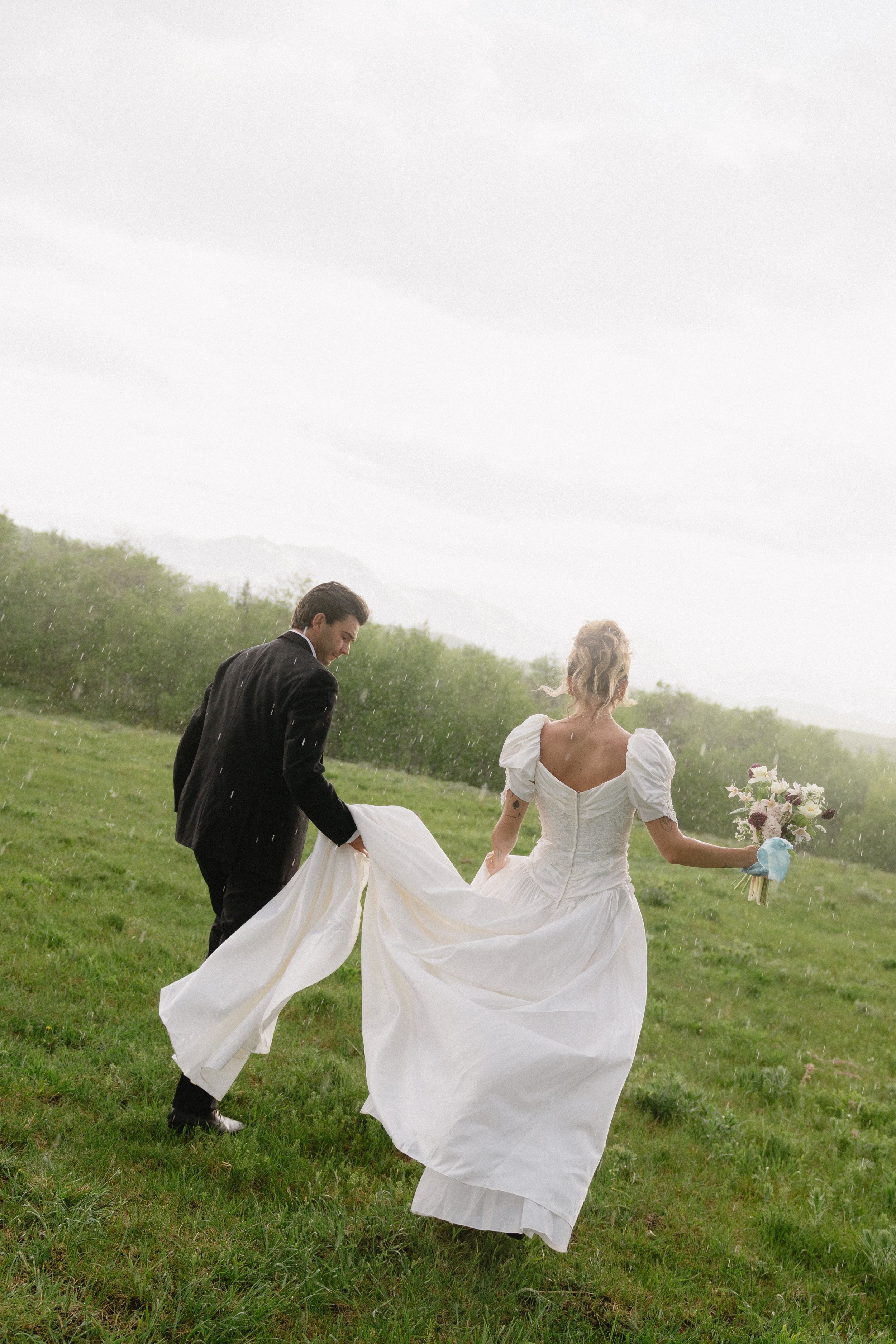 A bride and groom walking through a grassy field in rainy weather, with the groom lifting the bride's dress and holding her hand, as the bride carries a bouquet of flowers.