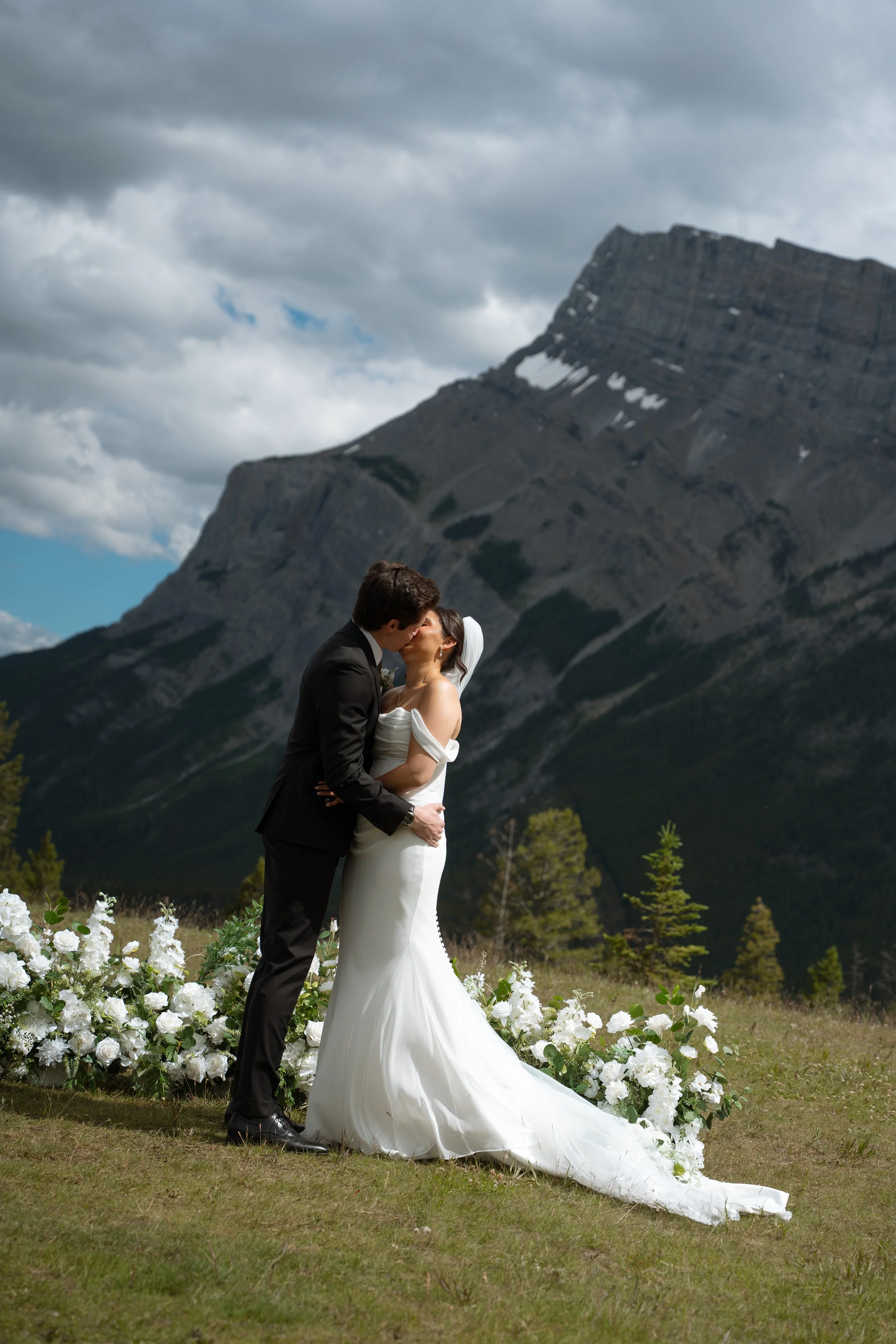 A couple in wedding attire sharing a kiss outdoors with mountains and cloudy sky in the background, surrounded by white flowers.