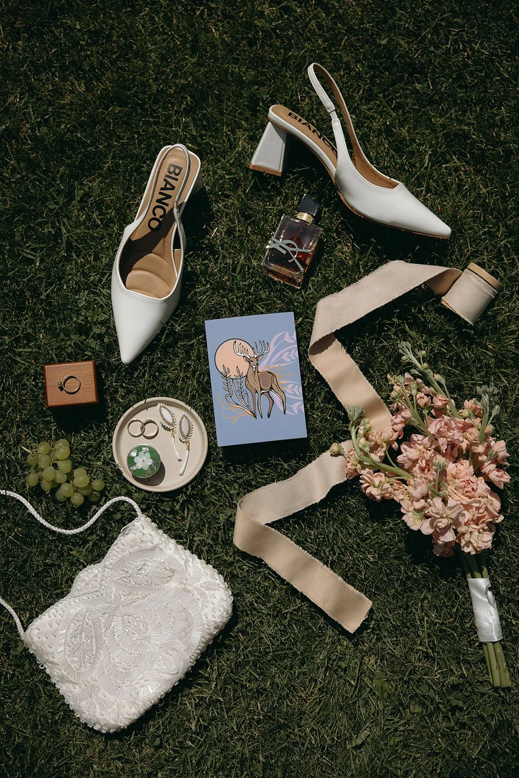 White high-heeled shoes, perfume bottle, bouquet of pink flowers, white lace bag, jewelry, postcard with a deer illustration, grape bunch, ribbon, ring, earrings, and a small box with a ring, scattered on grass.