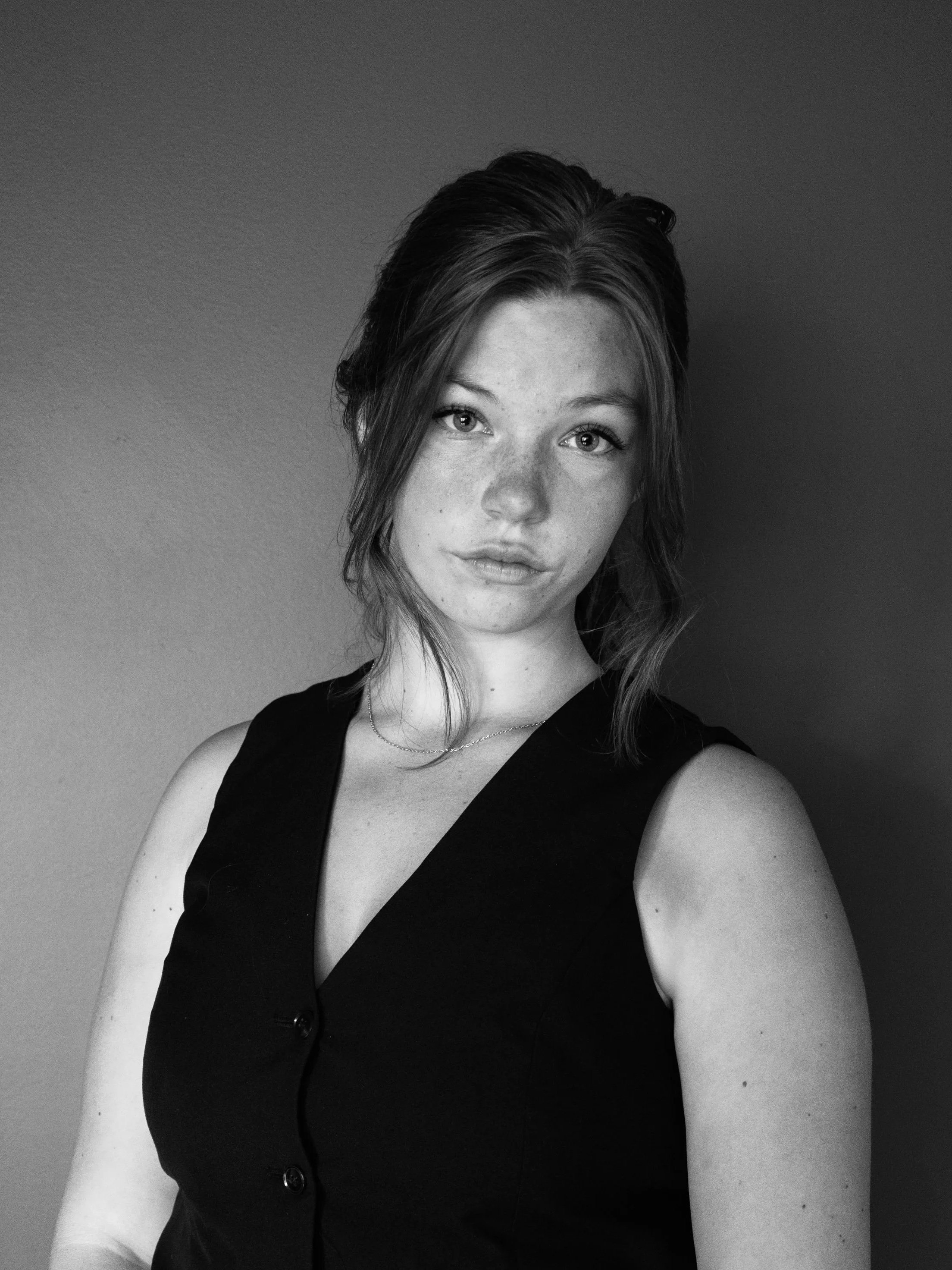 Black and white portrait of a woman with shoulder-length hair, wearing a sleeveless vest with buttons and a delicate necklace, looking directly at the camera against a plain background.