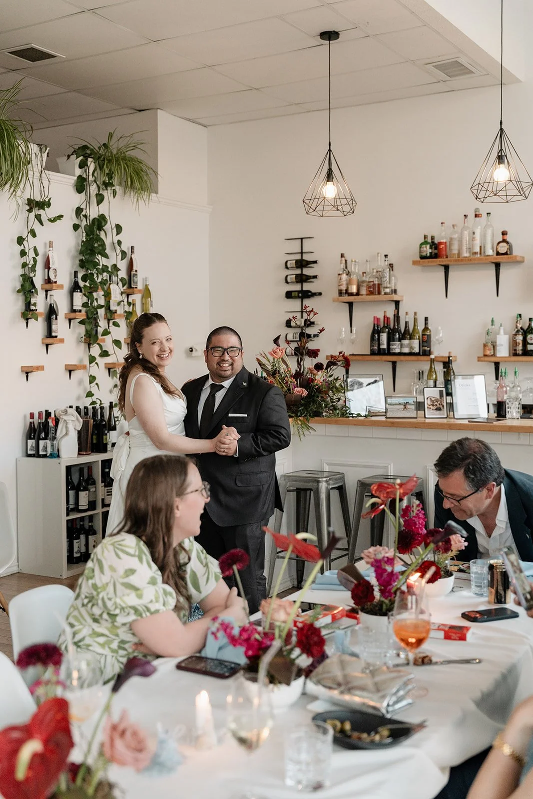 A couple is dancing at a wedding reception, with guests seated at a decorated table in the foreground.