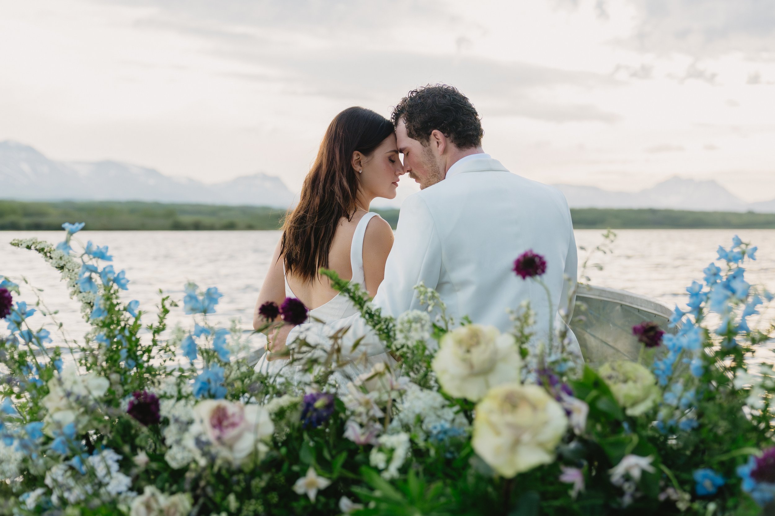A couple in wedding attire touching foreheads on a boat surrounded by colorful flowers with a water and mountain landscape in the background.