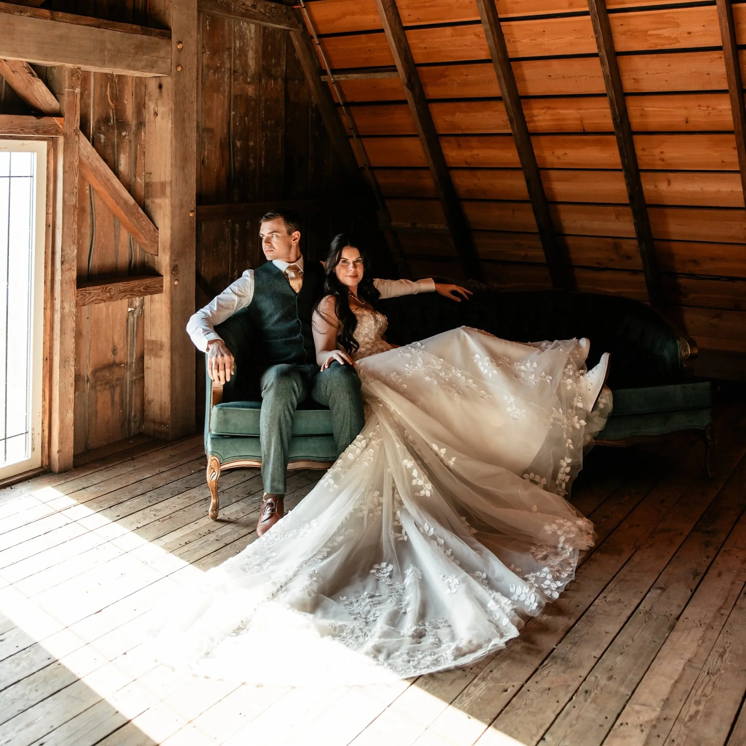 A man and woman in wedding attire sitting on a vintage green sofa in a rustic wooden room, with a high sloped wooden ceiling and a window letting in natural light.