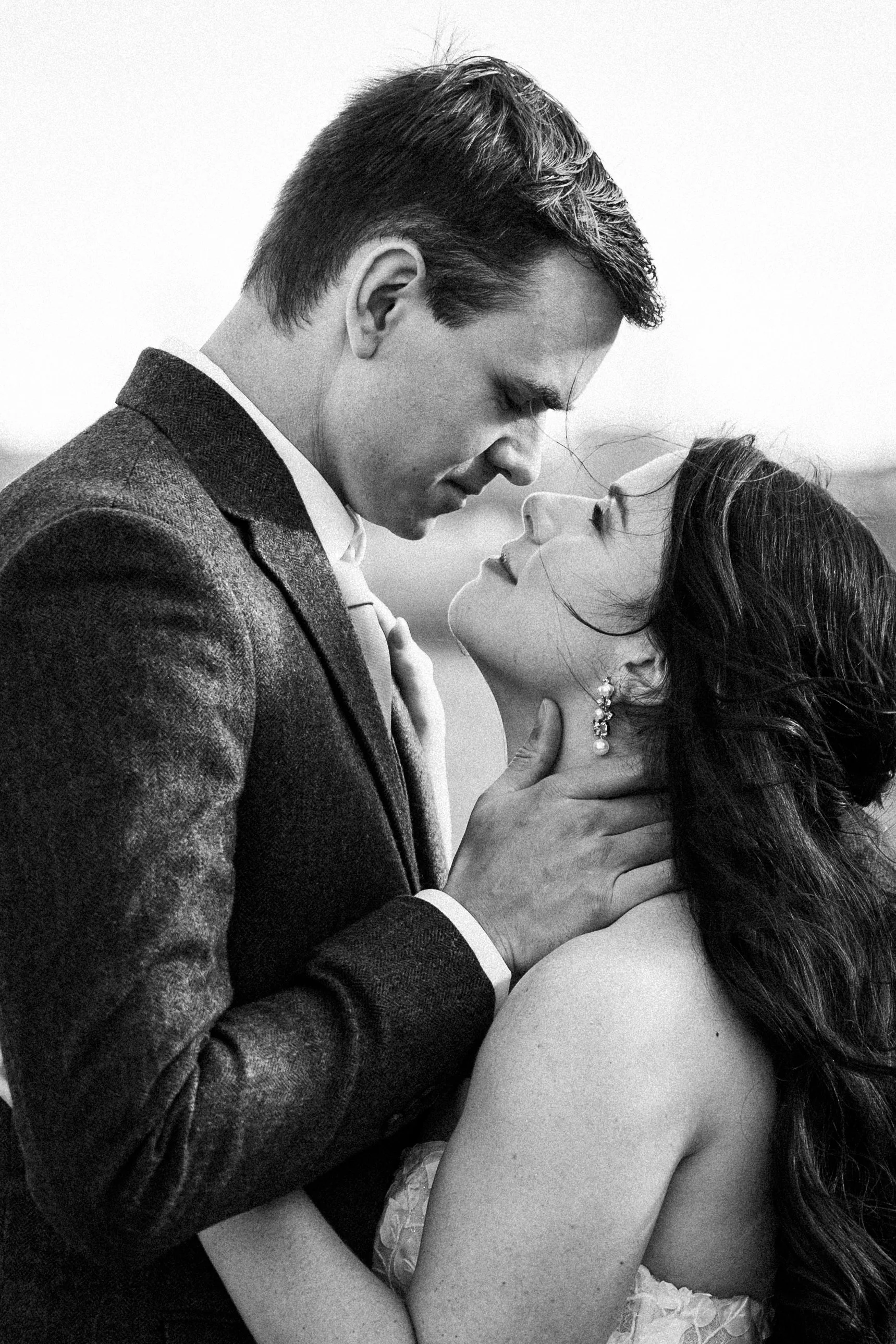 A black and white photo of a man and woman in a romantic embrace, about to kiss. The man is wearing a suit and the woman is in a dress, with her eyes closed and head tilted upward.