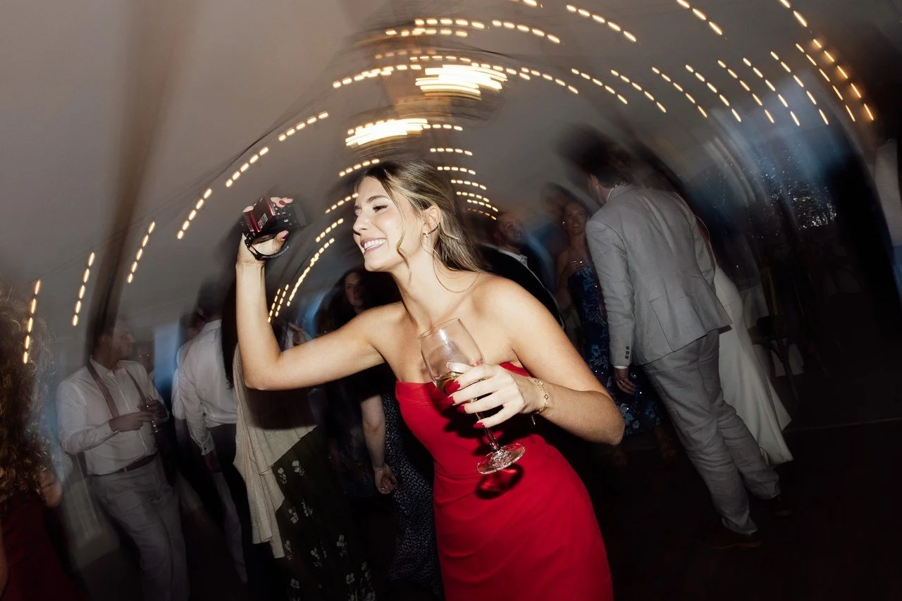 A woman in a red dress dancing and holding a drink at a lively party with other guests, some in formal attire, under a ceiling with string lights.