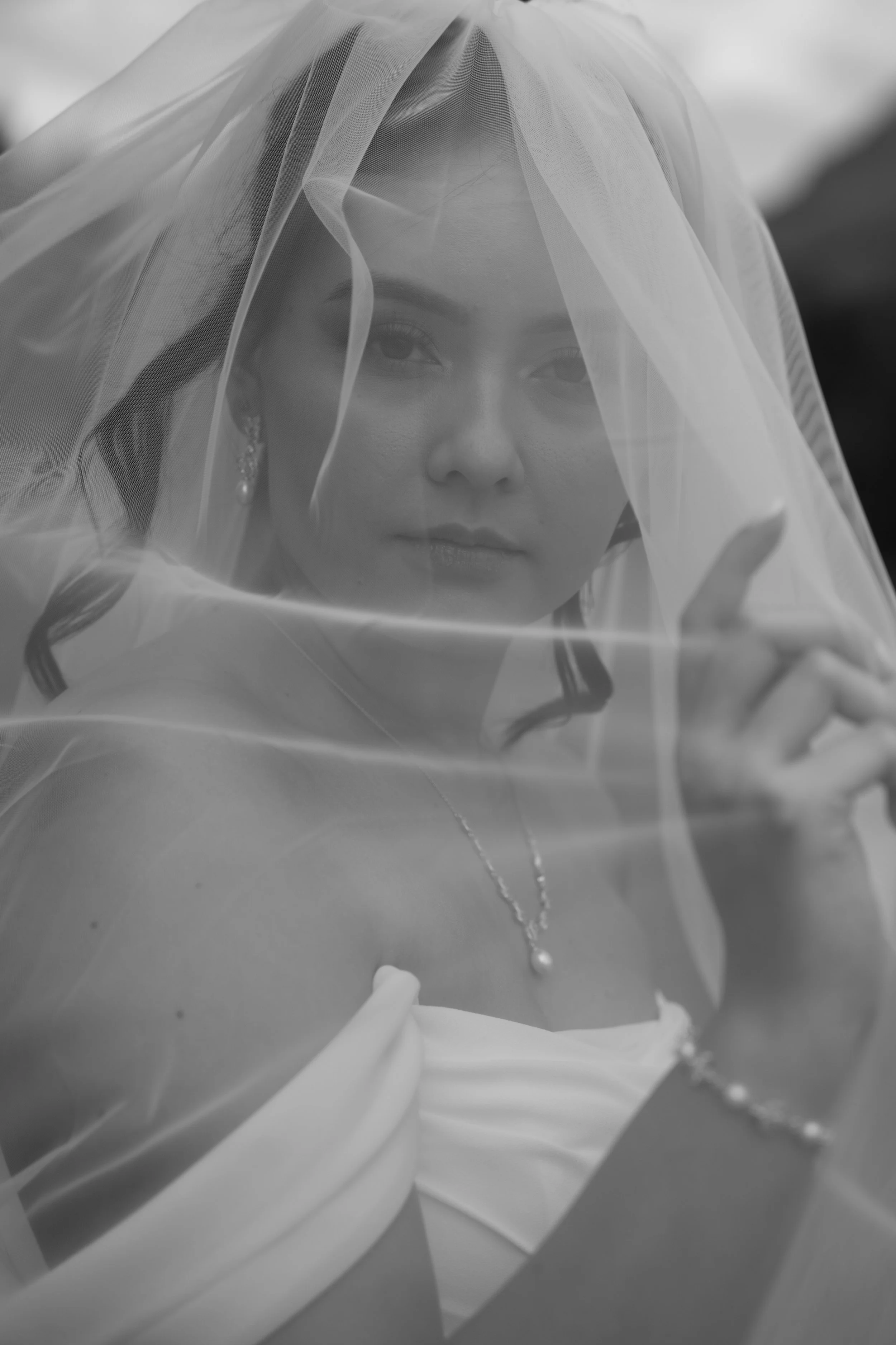 A black and white photo of a woman with a veil covering her head and shoulders, looking at the camera with a serene expression, wearing earrings and a necklace.