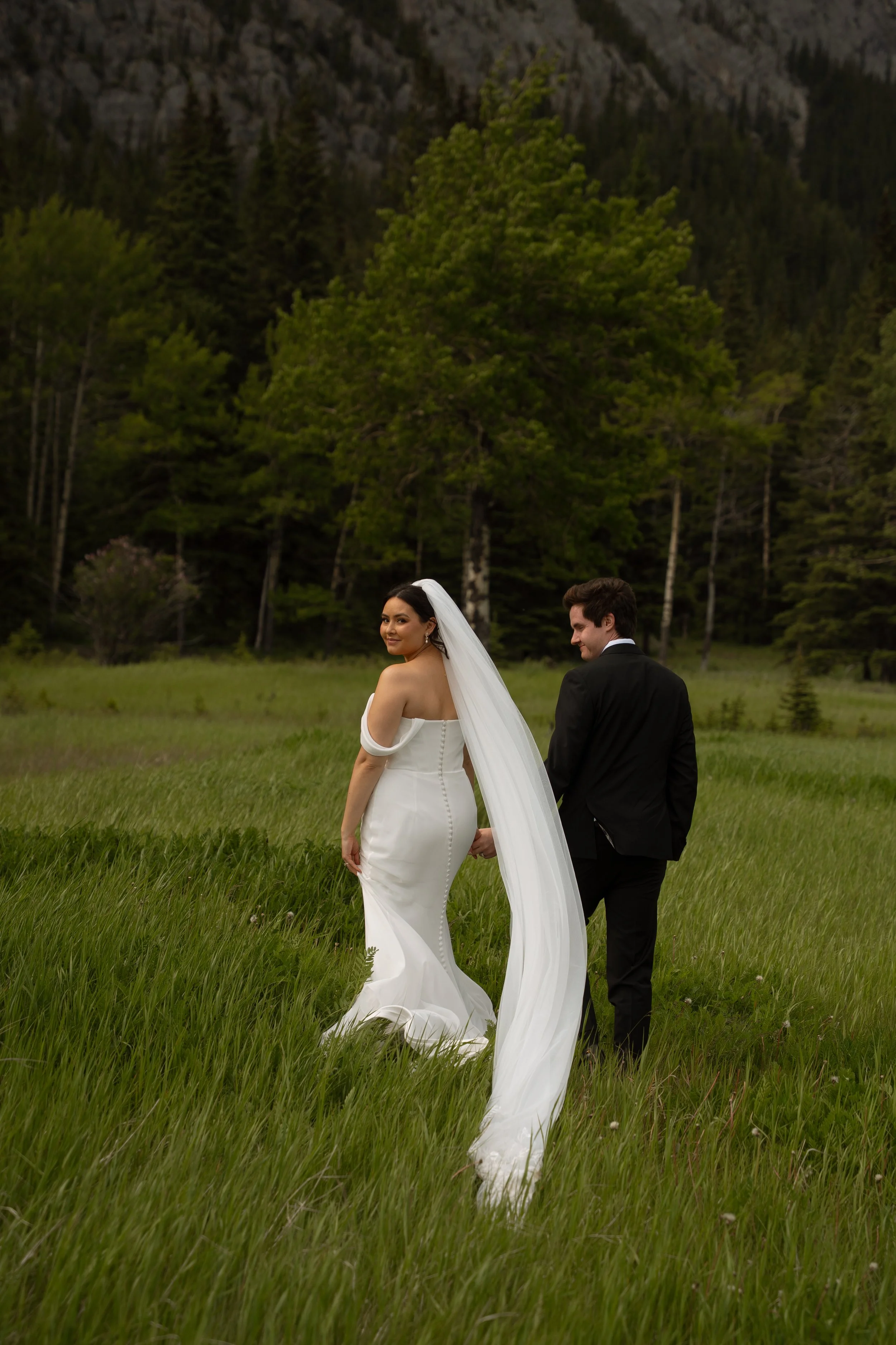 A bride and groom holding hands in a grassy field with a backdrop of trees and mountains.