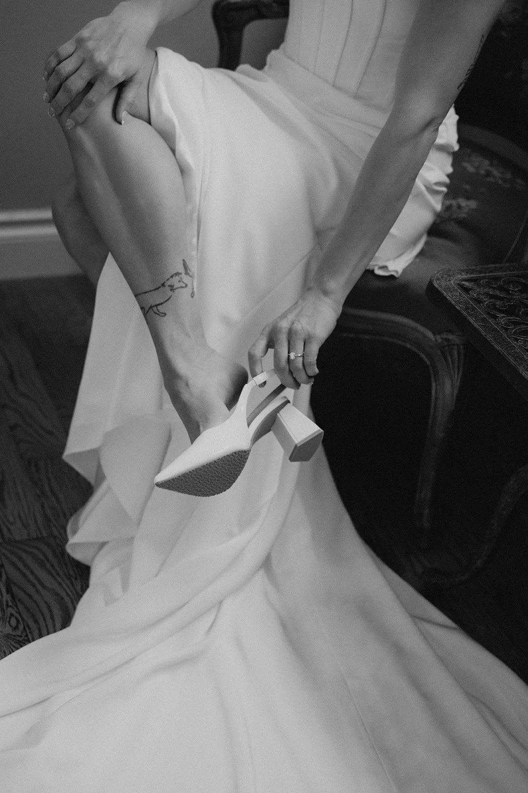 Person sitting on a chair, wearing a white dress, raising one leg, and holding a high-heeled shoe in their hand.