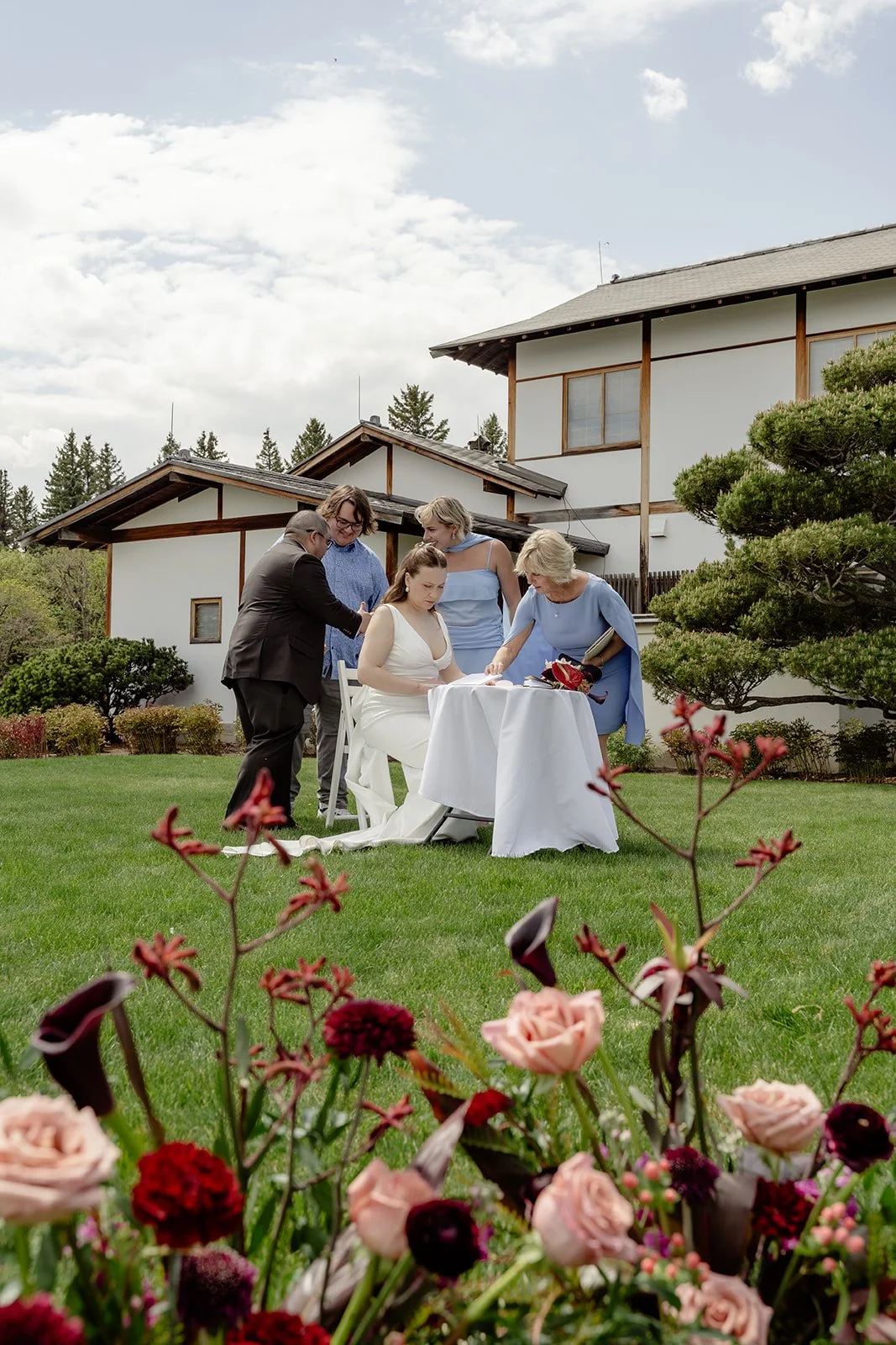 A wedding ceremony taking place on a grassy lawn with greenery, flowers, and trees, outside a large house or lodge, with five people gathered around a woman in a white wedding dress sitting at a small table.