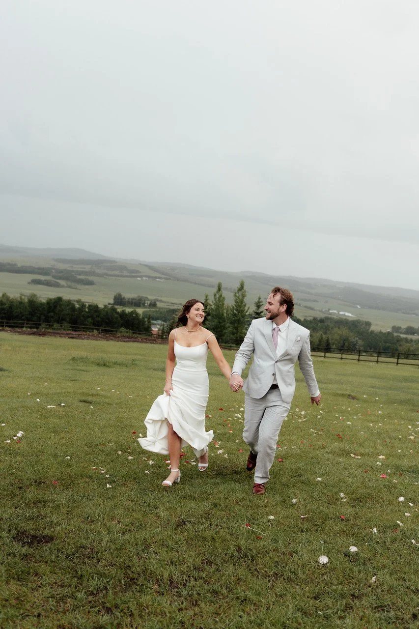 A bride and groom walking hand in hand across a grassy field on their wedding day, with a cloudy sky and distant hills in the background.