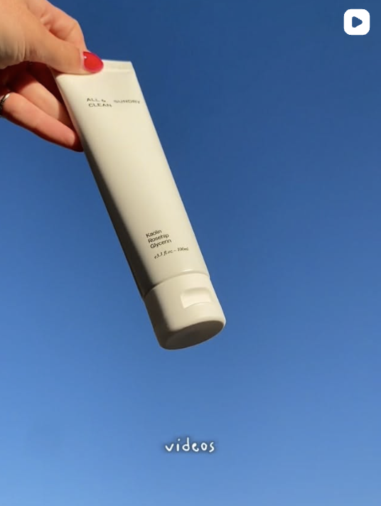 A hand holding a white tube of skincare product against a clear blue sky.