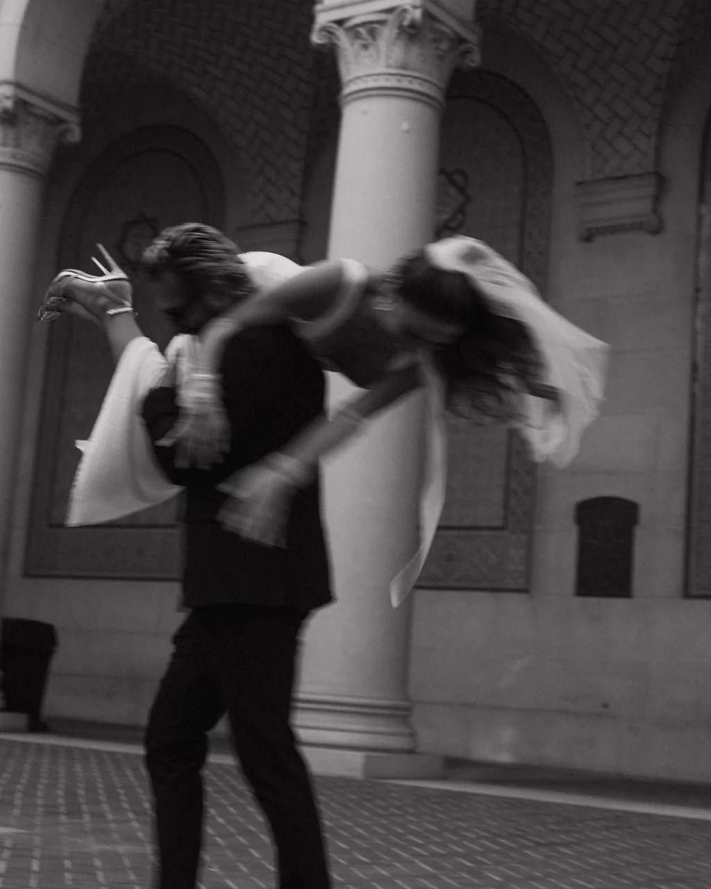 A black and white photograph showing a man carrying a woman over his shoulder inside a building with classical architectural elements.