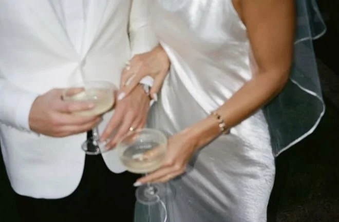 Close-up of a couple holding champagne glasses, dressed in white and silver, celebrating at a wedding or special event.