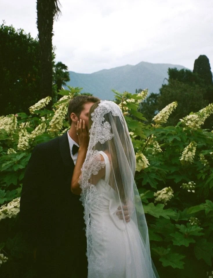 A bride and groom share a kiss outdoors with lush green foliage and white flowers, mountains in the background.
