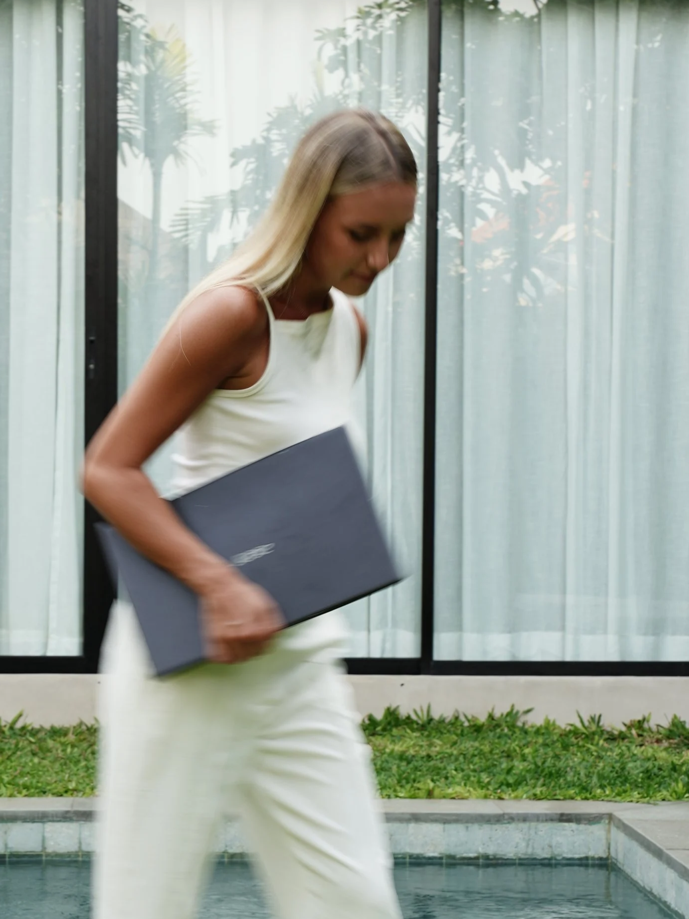 A woman with blonde hair wearing a white sleeveless top and white pants, holding a black folder, walking past a large window with curtains and greenery outside.
