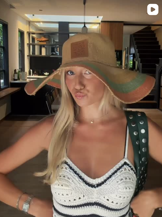 A woman wearing a large beige sun hat with a green and orange stripe, making a playful facial expression, standing inside a modern house with open kitchen and stairway in the background.